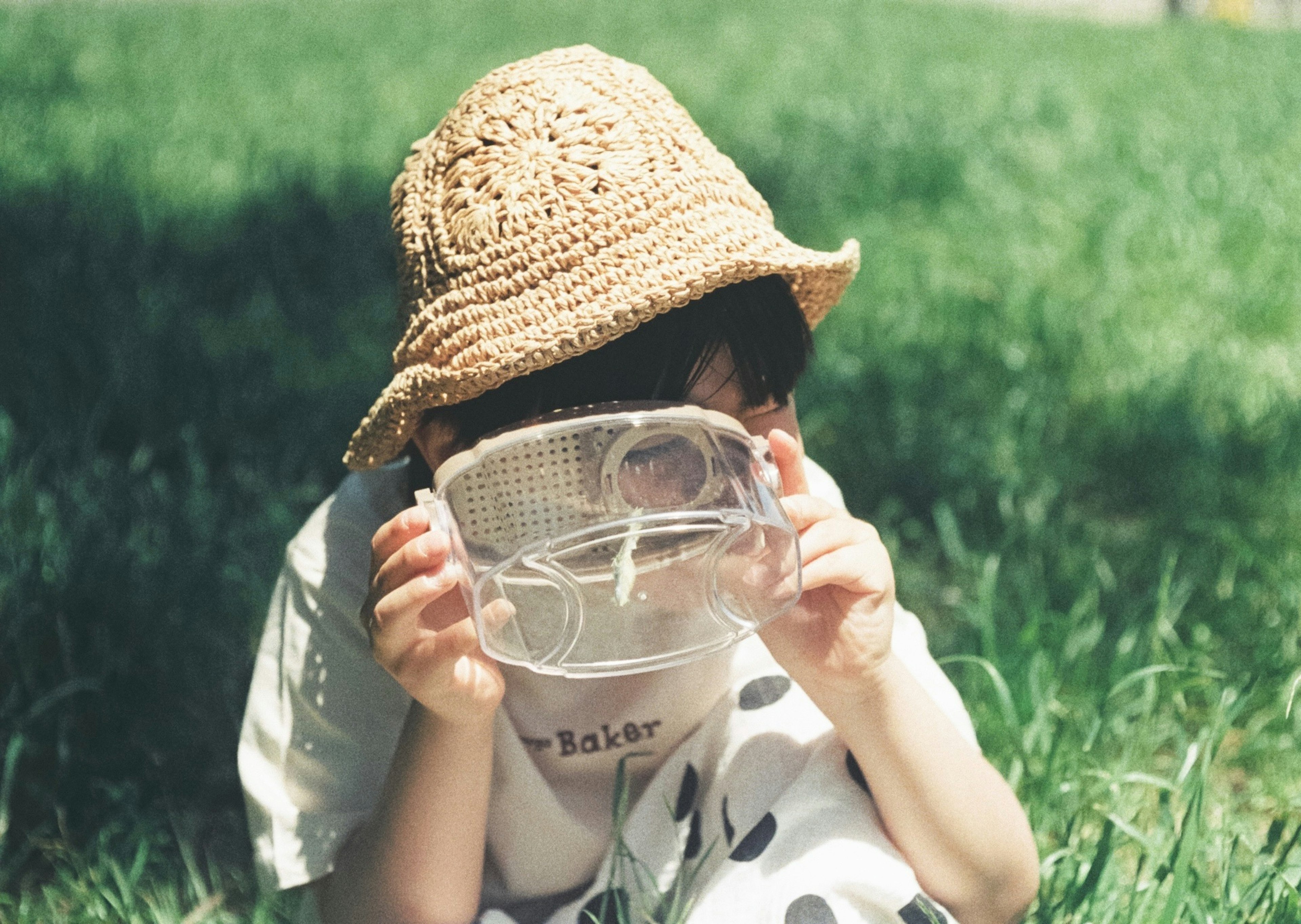 Bambino con cappello di paglia seduto sull'erba che tiene una maschera trasparente