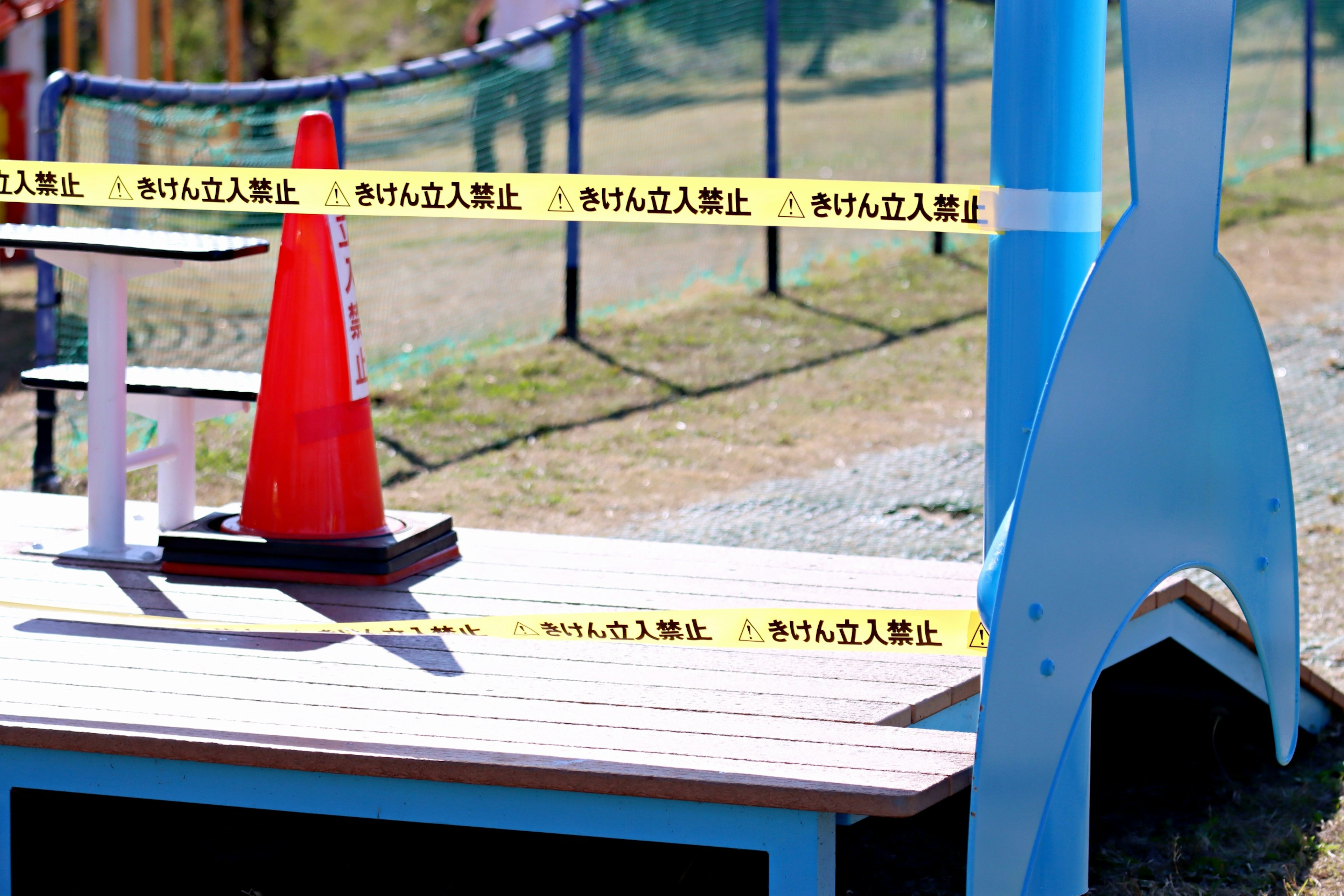 Área del parque con equipo de juego azul y un cono rojo rodeado de cinta amarilla de advertencia