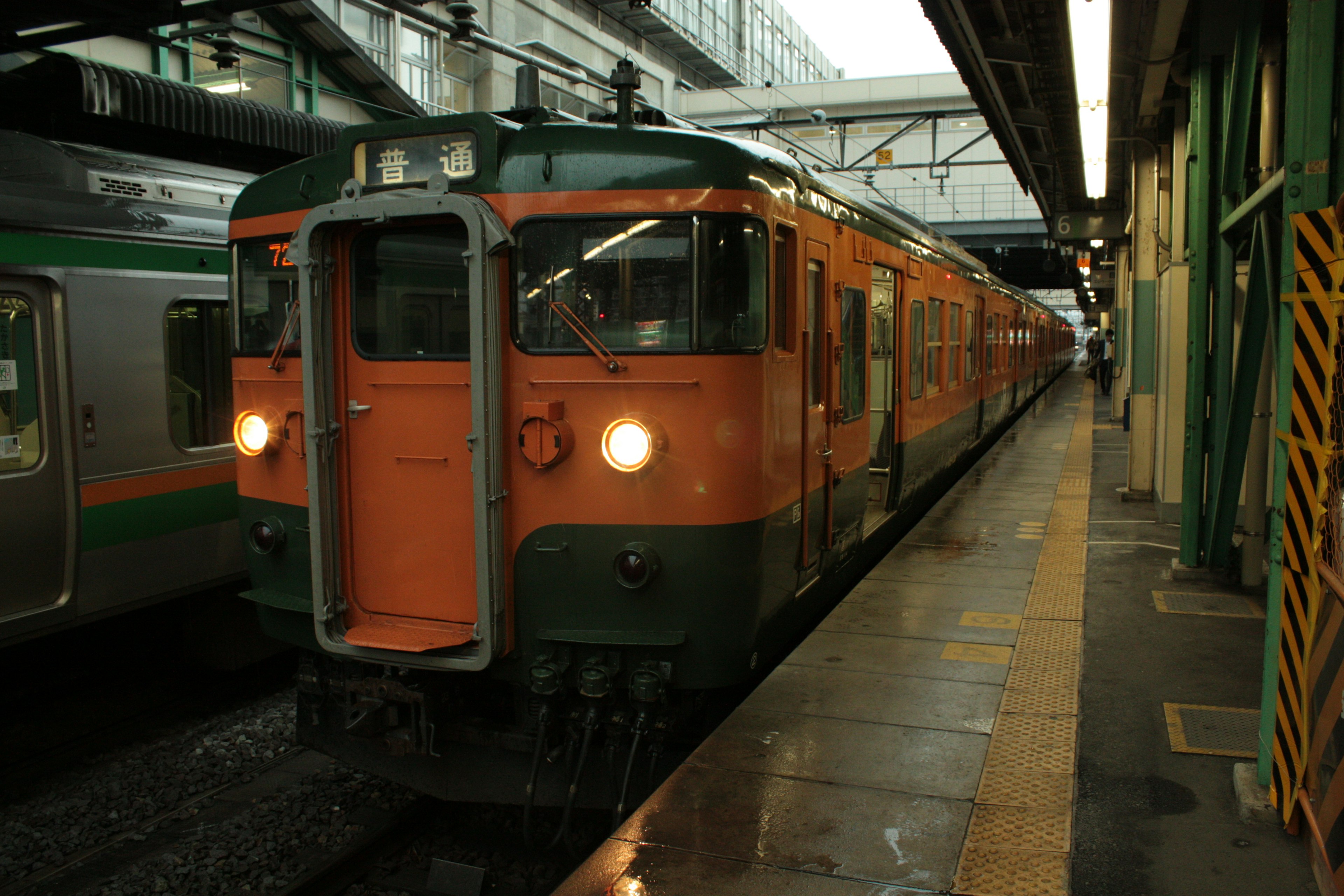 Ein orangefarbener Zug hält an einem Bahnhof mit anderen Zügen und einer sichtbaren Plattform