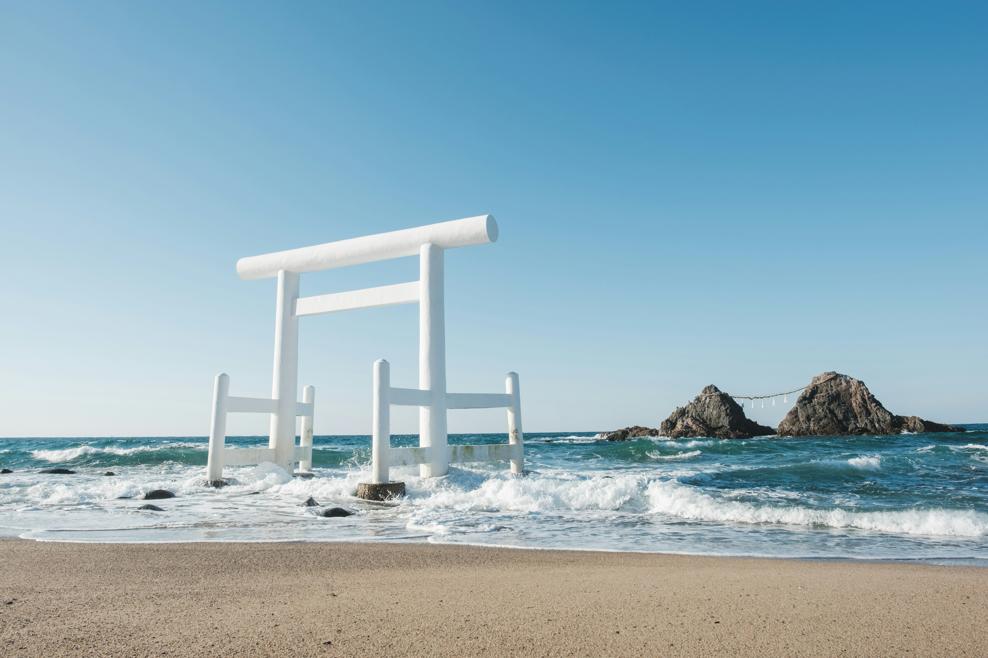 White torii gate stands on the beach with waves crashing