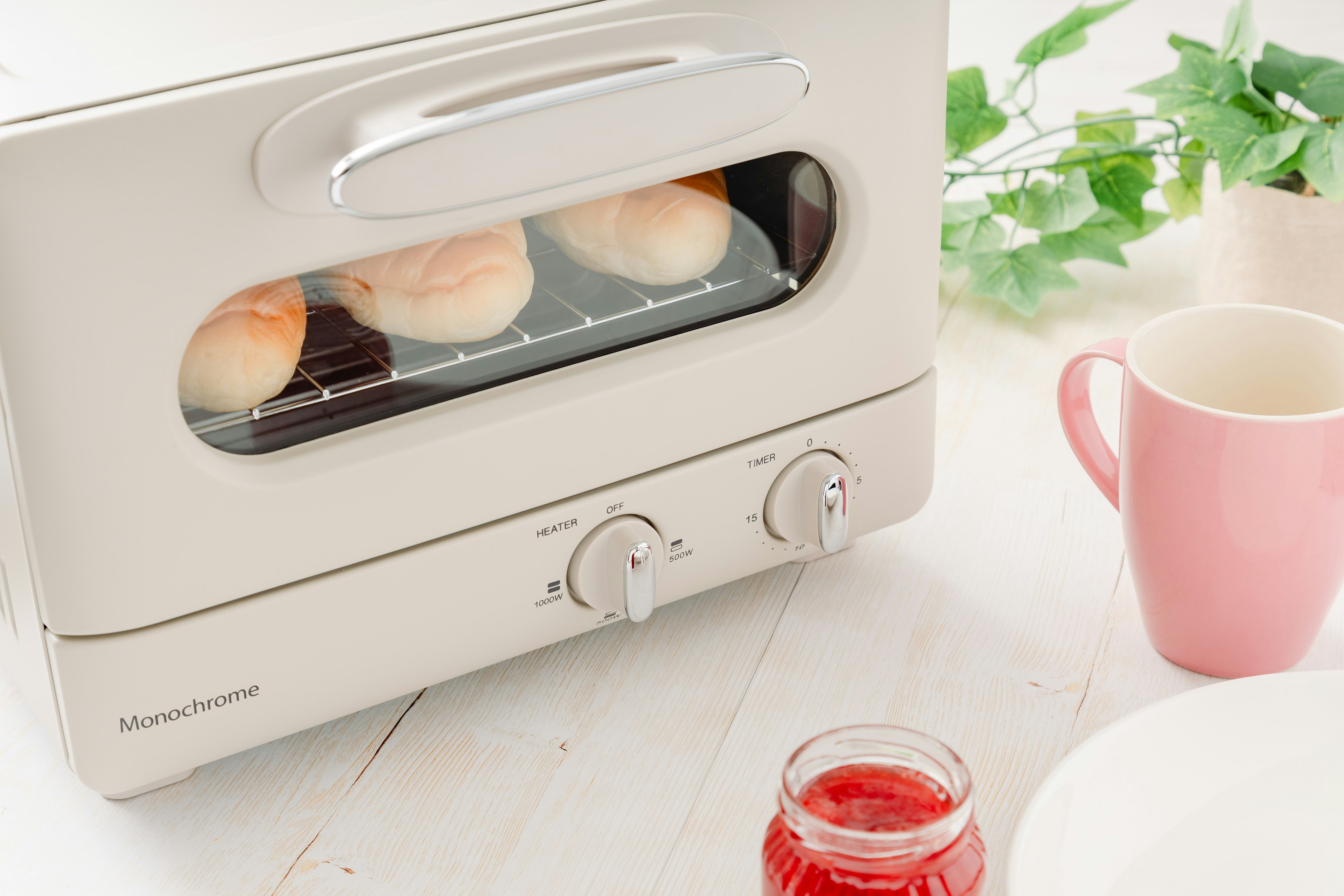 Một chiếc toaster màu trắng với bánh pancake bên trong và một chiếc cốc màu hồng bên cạnh một lọ mứt