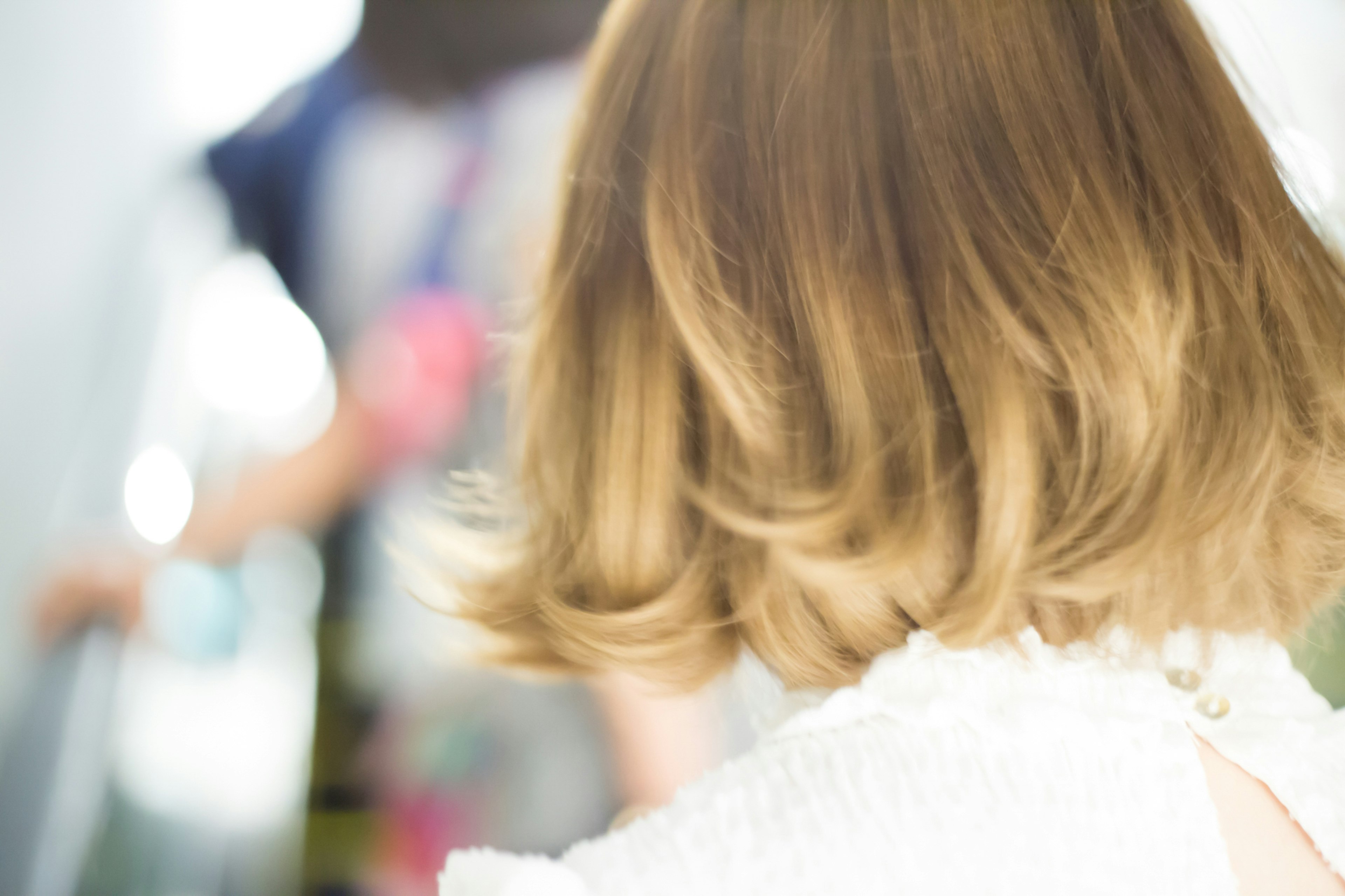 Close-up of a woman's blonde hair from the back with a blurred figure in the background