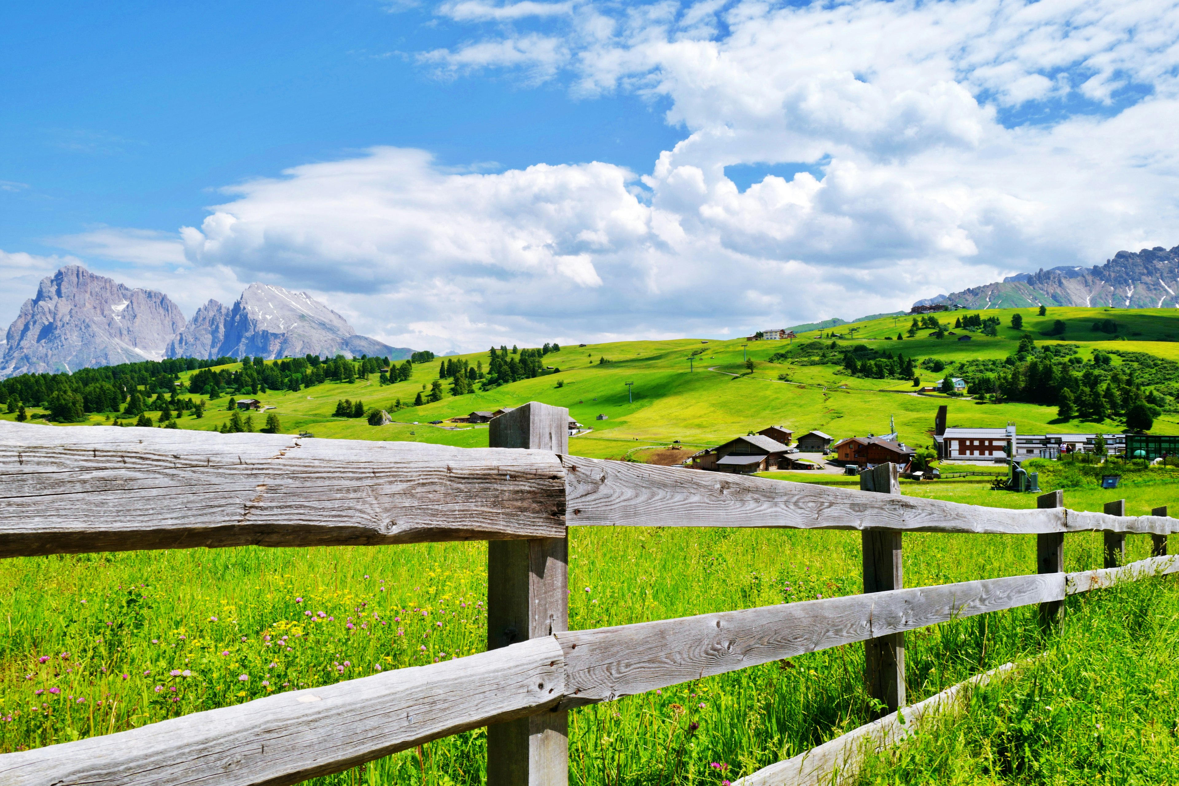 青い空と緑の牧草地が広がる風景 木製のフェンスが前景にあり 背景に山と家々が見える