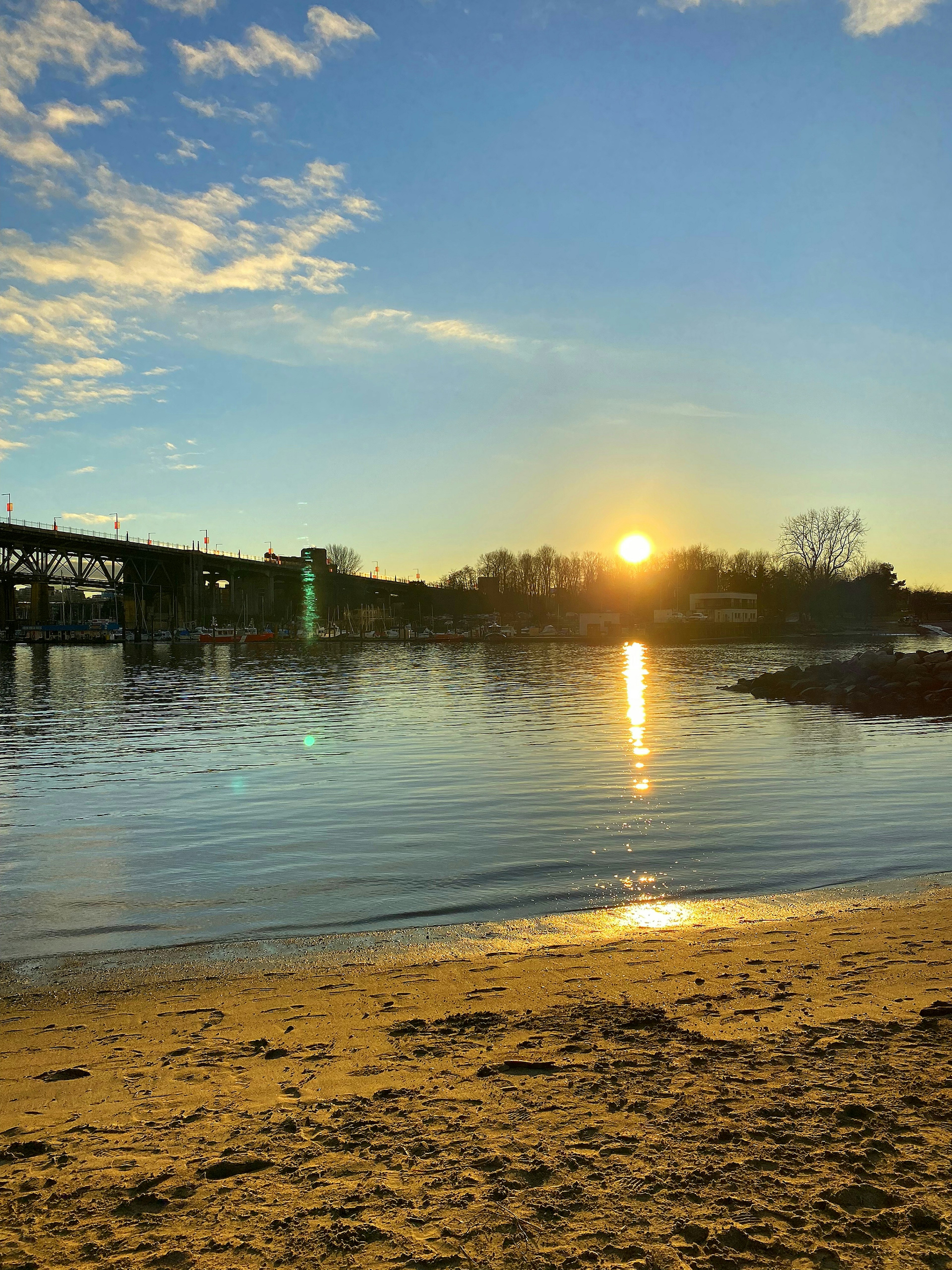 Pemandangan sungai dengan matahari terbenam yang memantul di air pantai berpasir dan jembatan yang terlihat