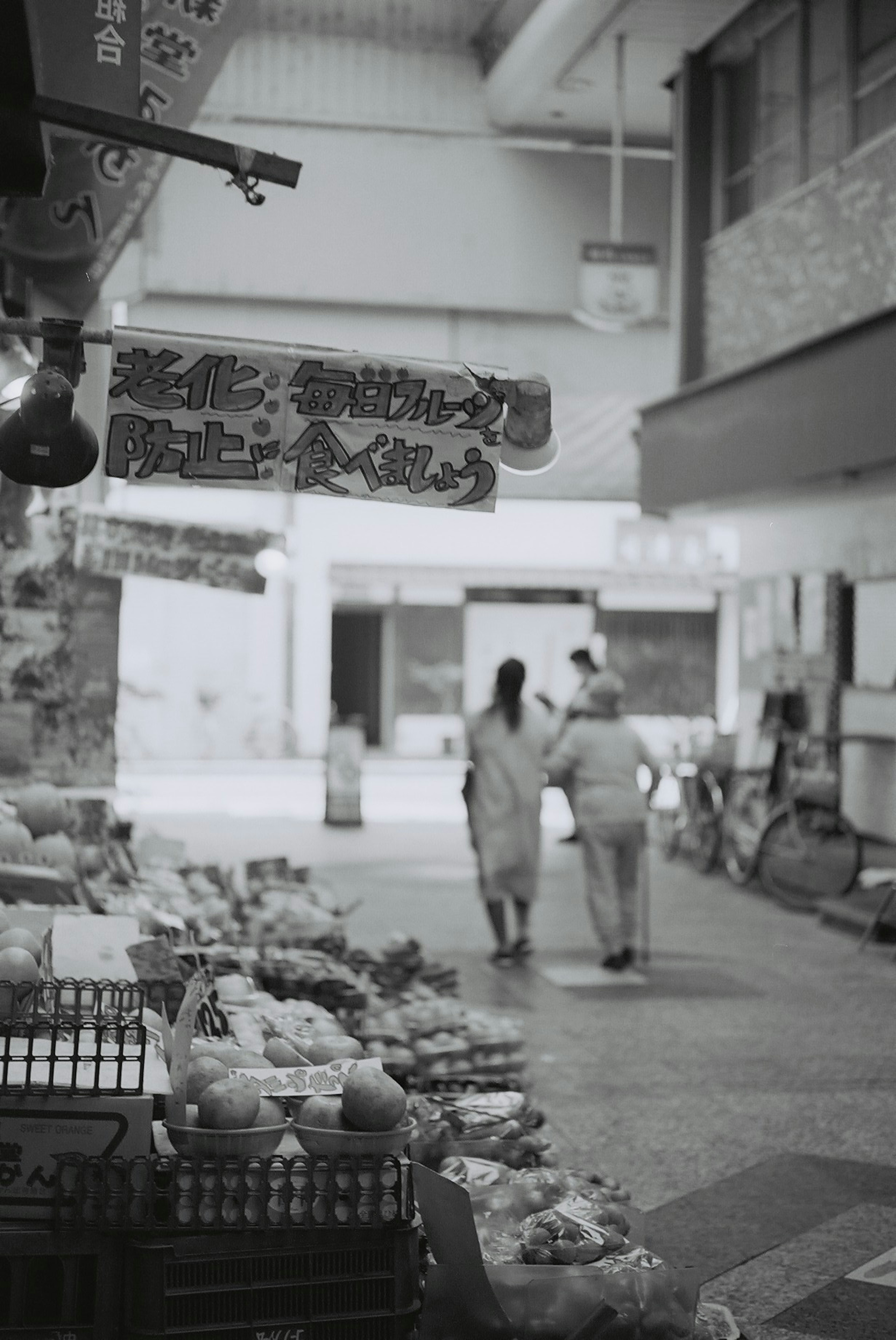 Deux femmes marchant dans une rue de marché en noir et blanc avec des étalages de légumes