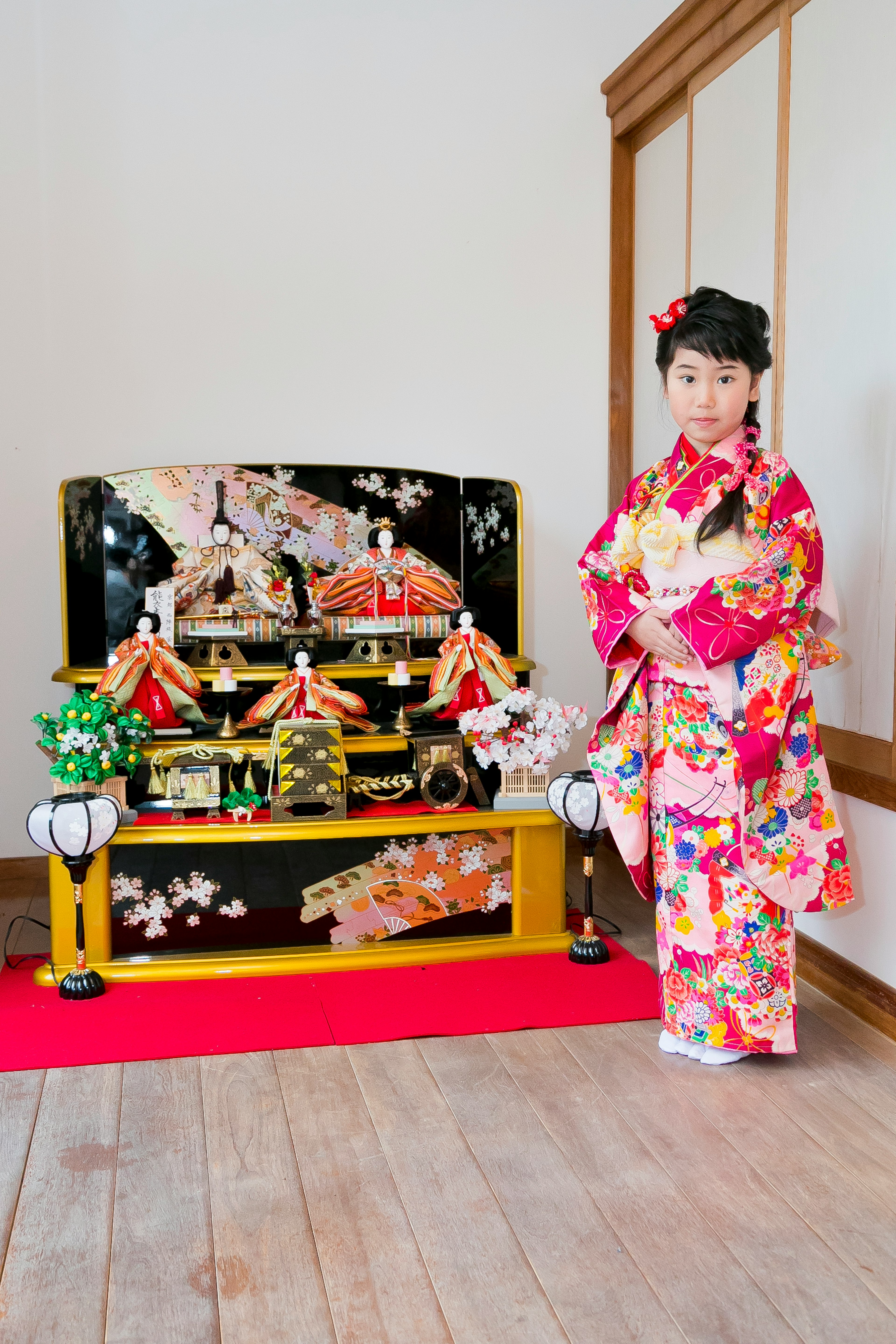 A girl in a colorful kimono stands in front of a hina doll display representing Japanese Girls' Day