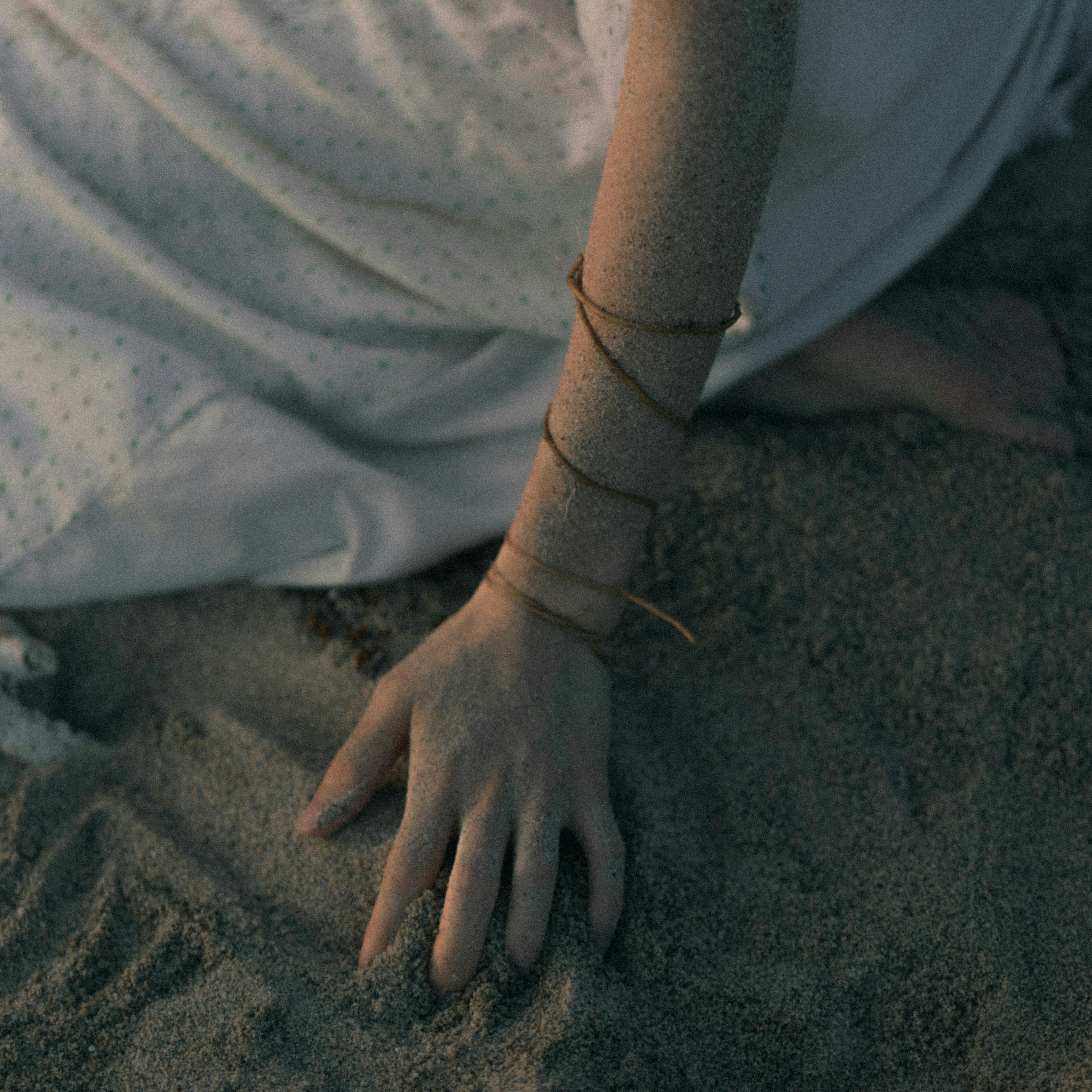 Eine Hand, die auf Sand ruht, mit einer Schnur um das Handgelenk einer Person in einem weißen Kleidungsstück