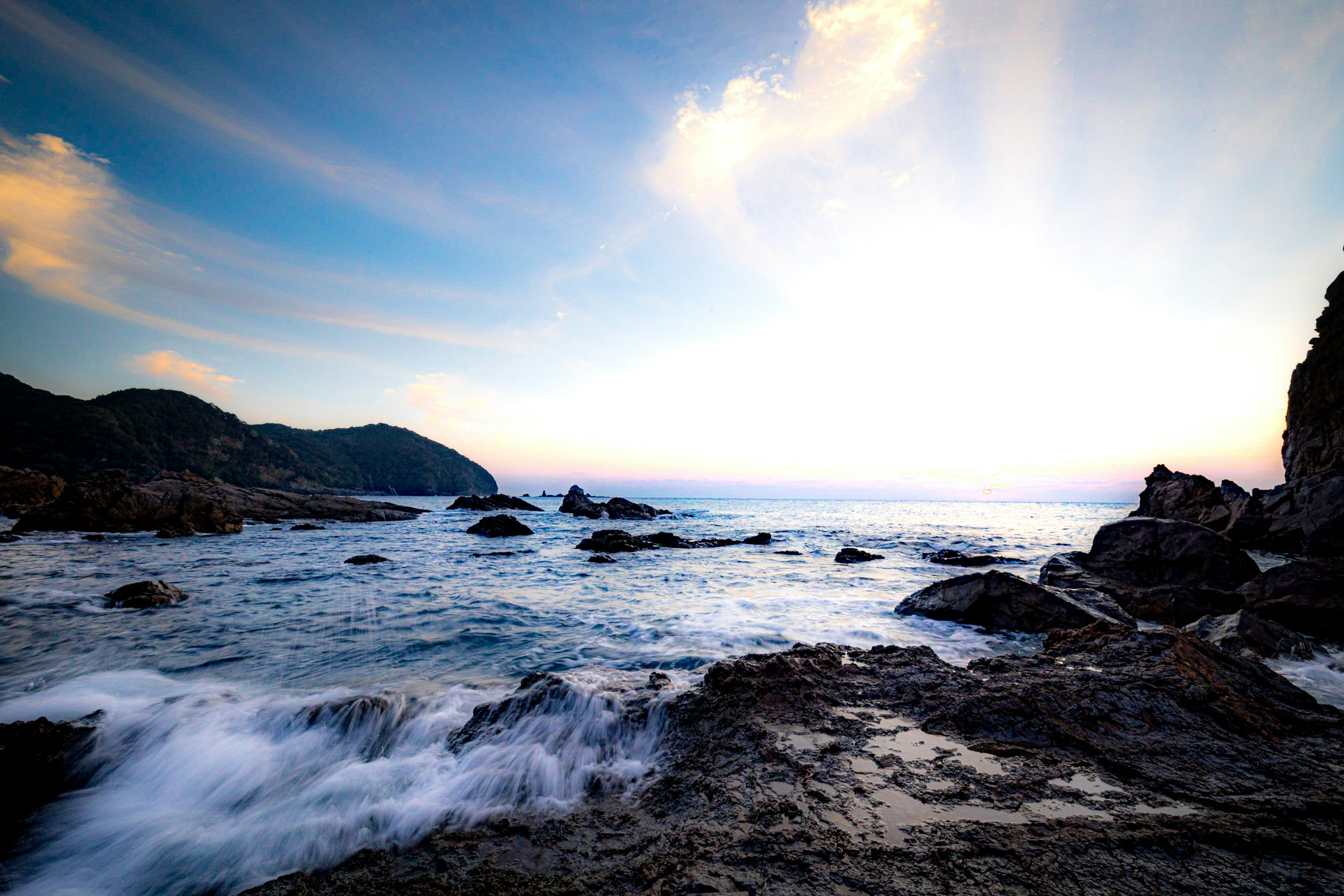 Pemandangan pantai dengan ombak menghantam batu, langit matahari terbenam yang indah dengan nuansa biru dan awan