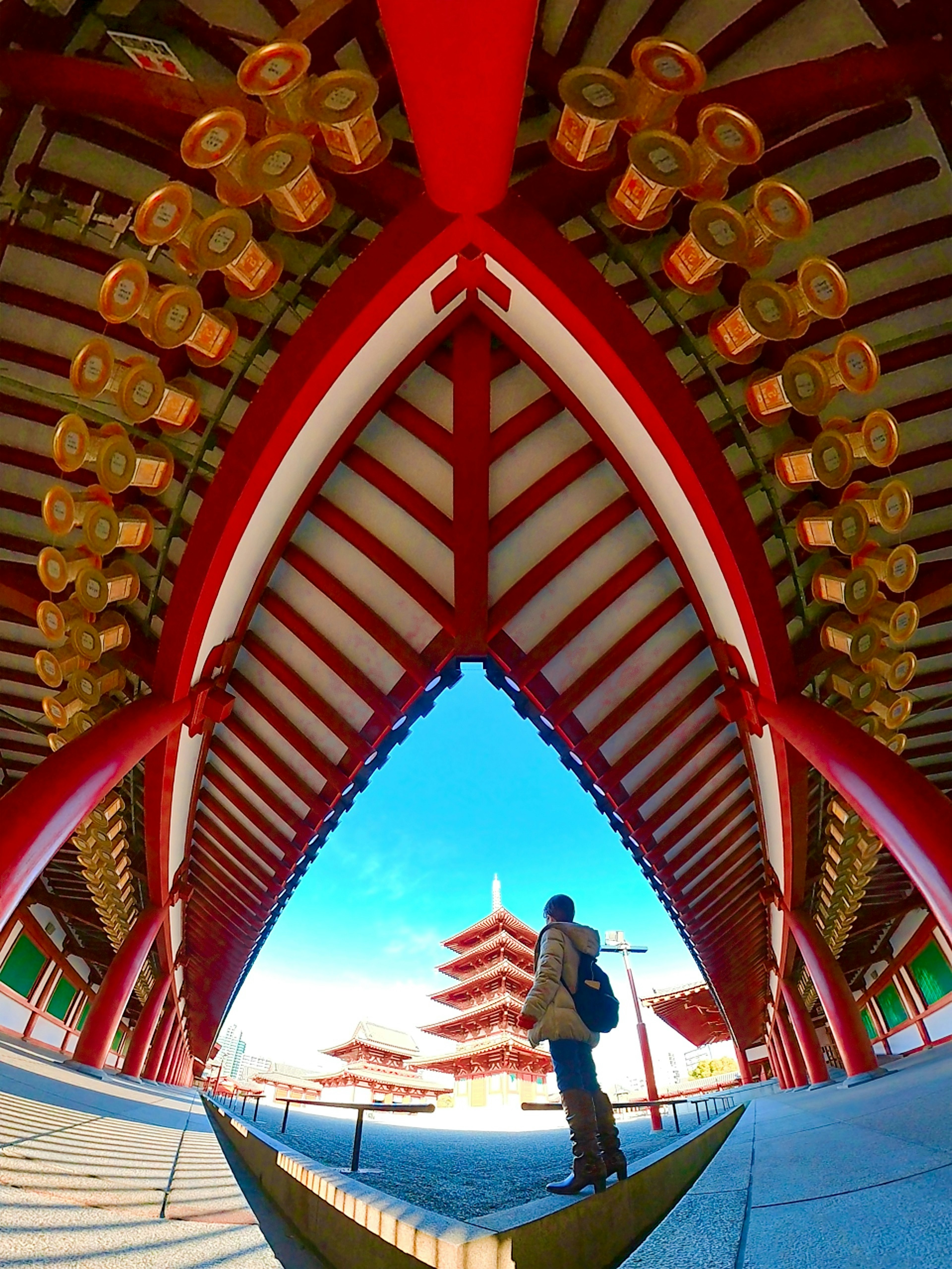 A person standing under an arch with red roof and large lanterns at a temple