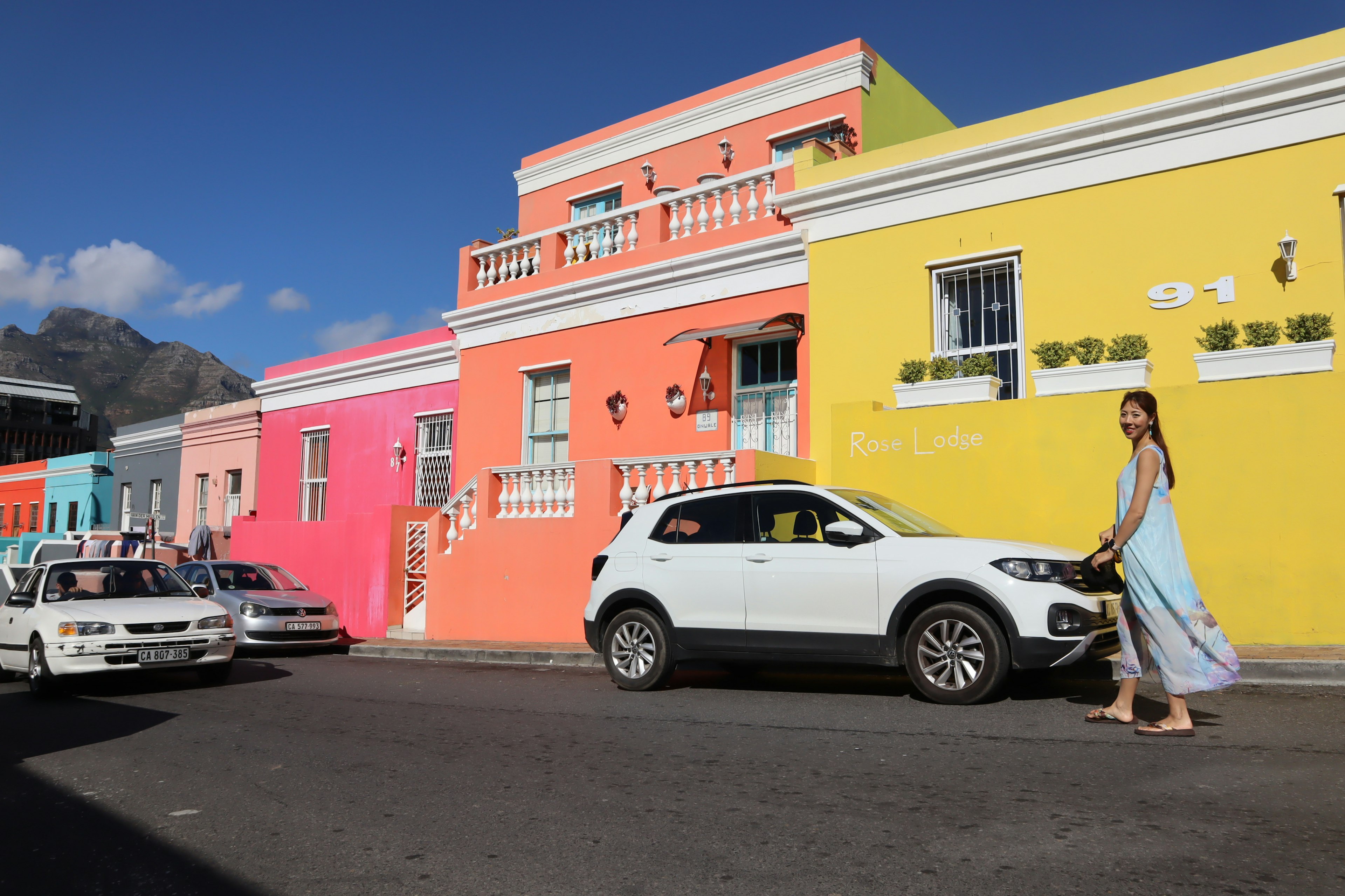 A woman walking along a colorful street with pink and yellow houses a white car and a blue sky