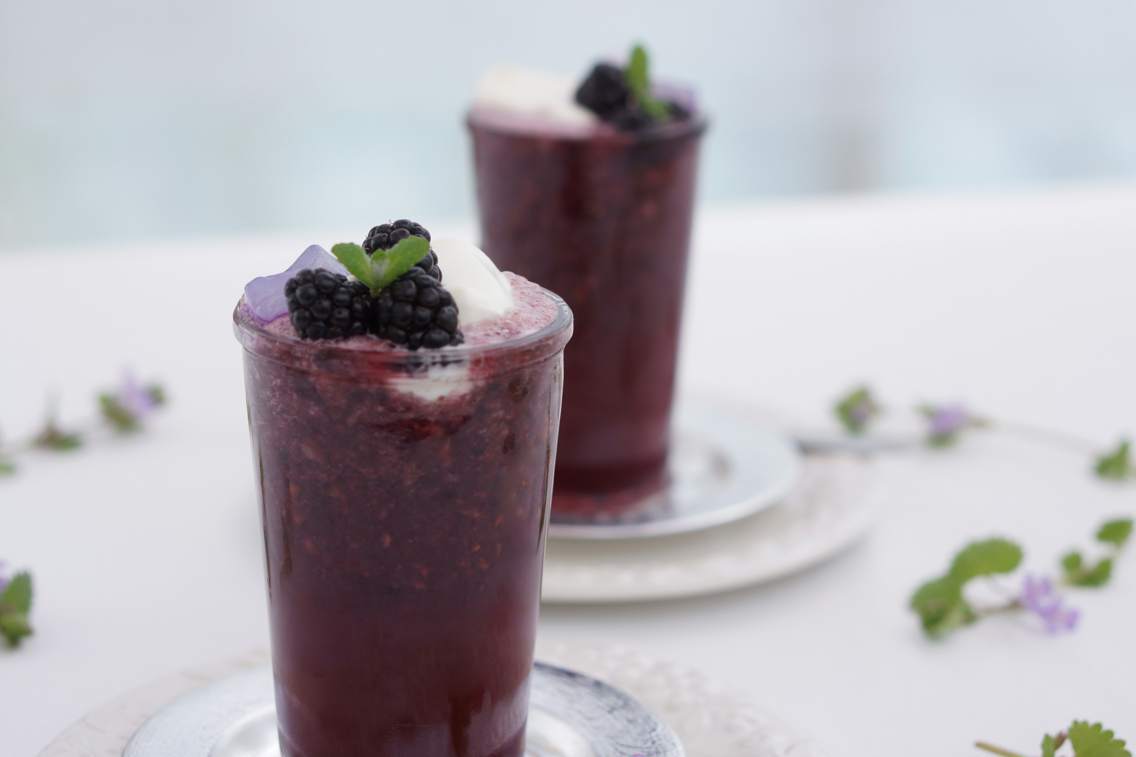 Two glasses of purple beverage topped with blackberries and mint on a table