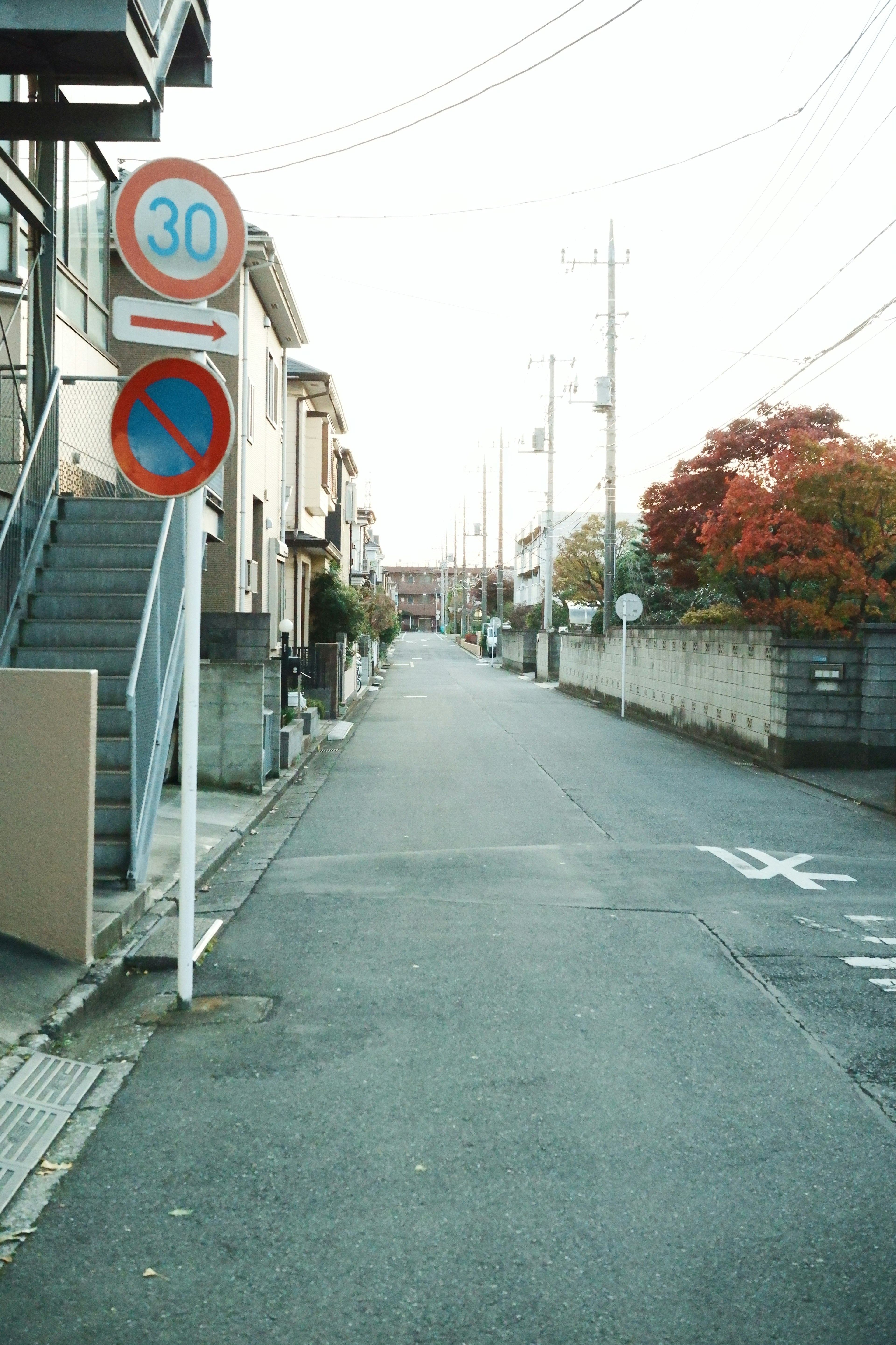 Enge Straße mit Wohngebäuden und einem Baum mit roten Blättern im Hintergrund