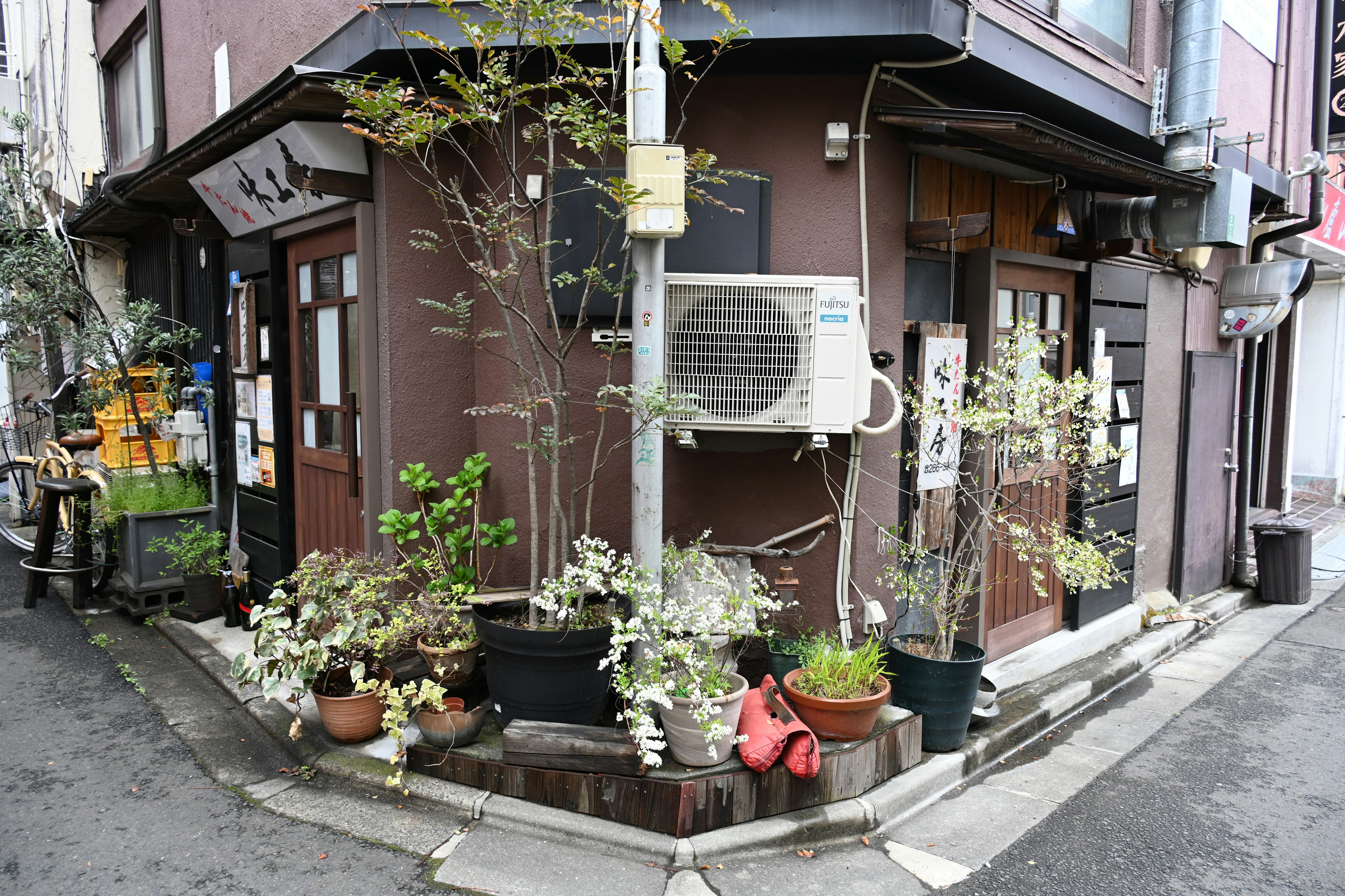 Small brown building at a street corner with lush green plants