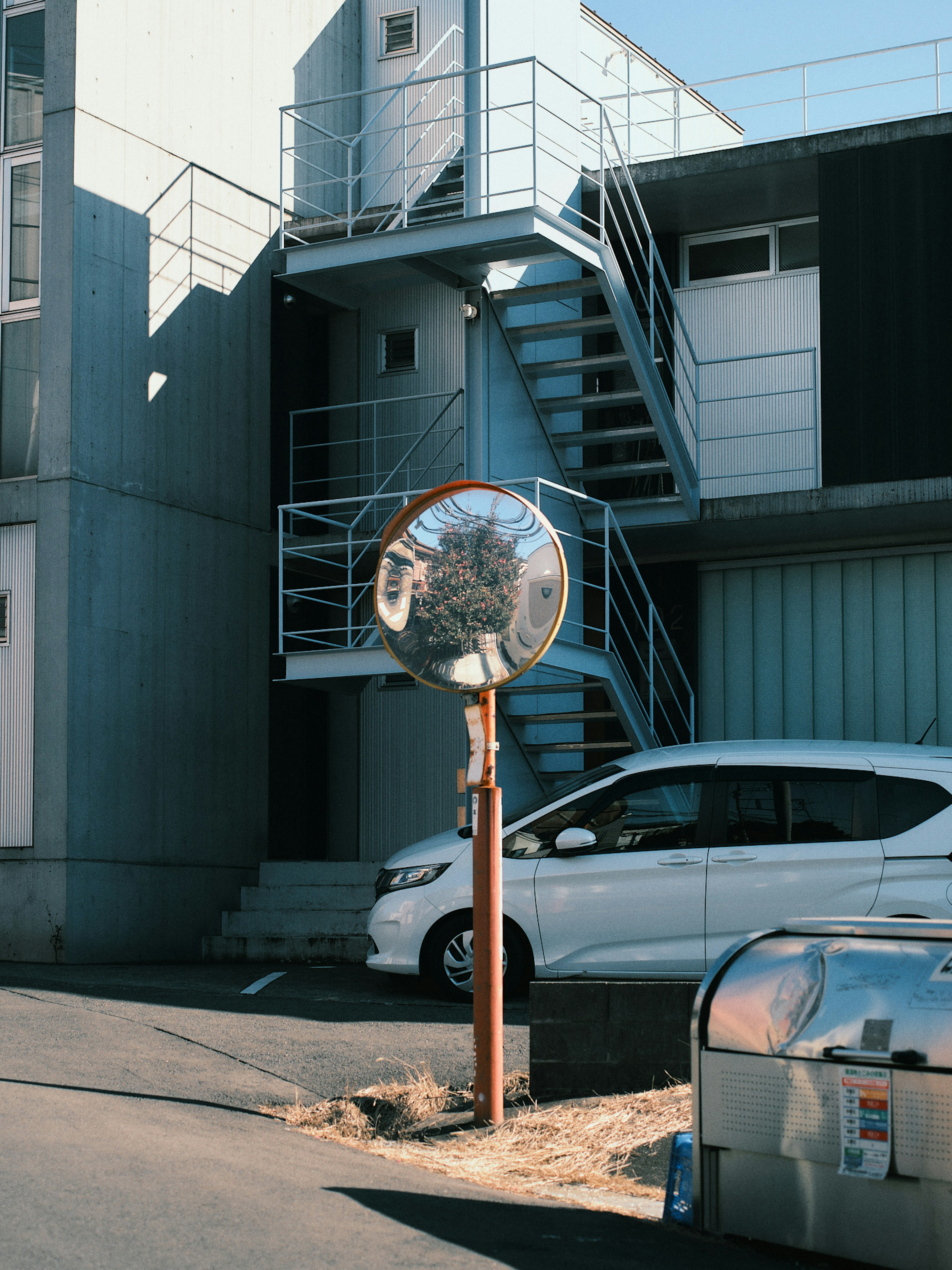 Vista exterior de un edificio con escaleras y un espejo que presenta un coche blanco y un cubo de basura de metal