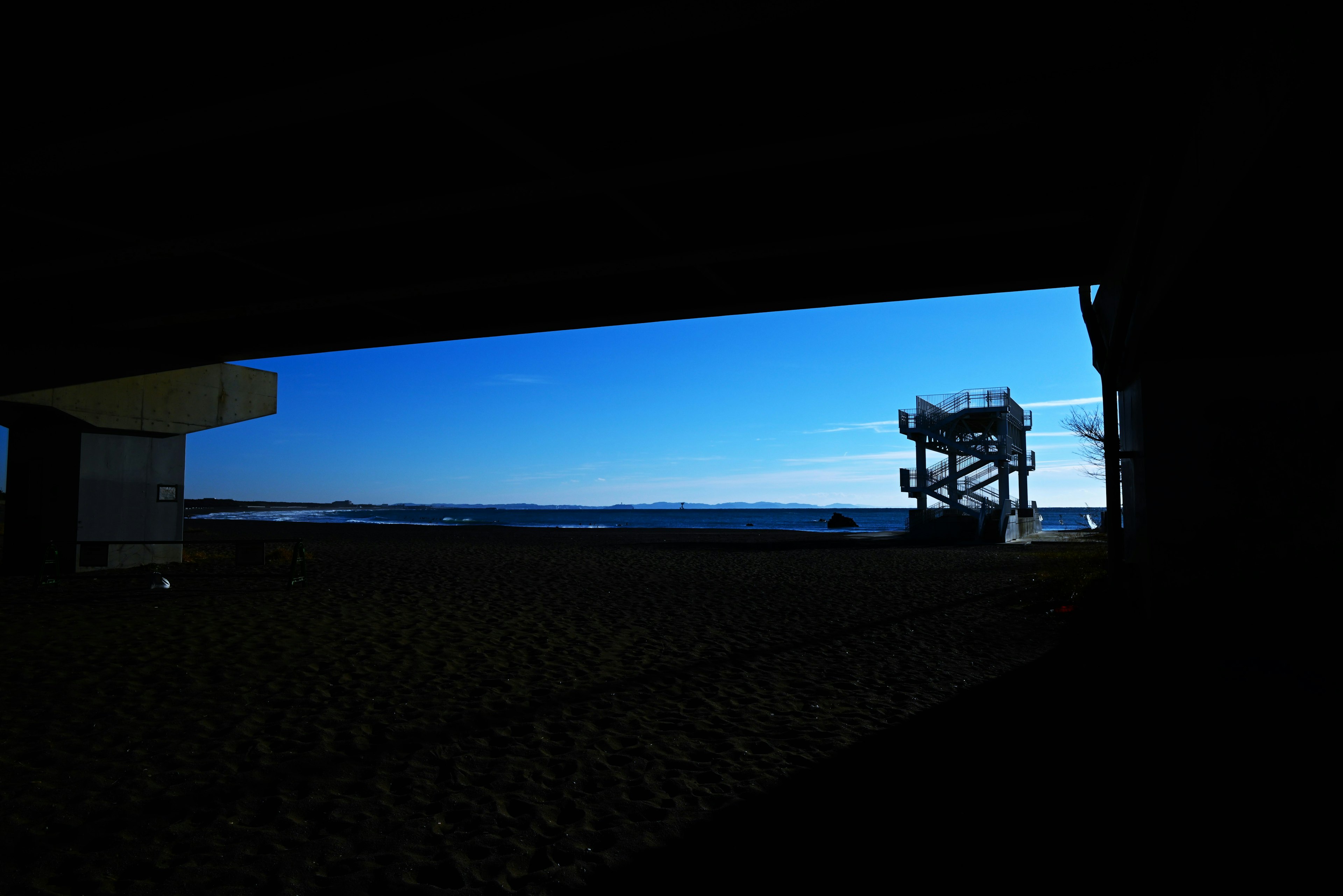 Vue d'une scène de plage avec un ciel bleu et un océan en arrière-plan, présentant un bâtiment à gauche et un poste de secours à droite