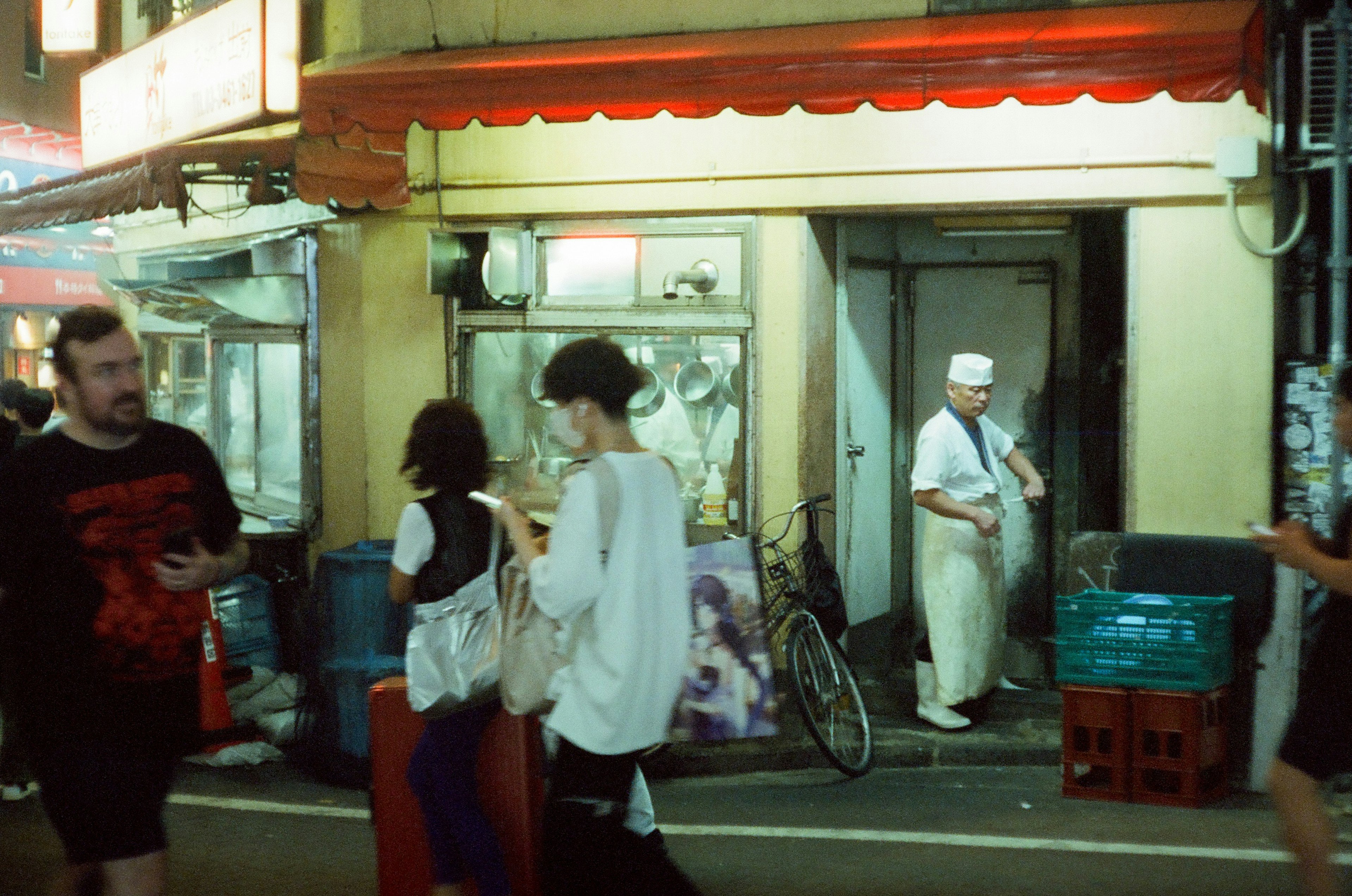 夜の街角で通行人が歩く中、白い帽子をかぶった老舗の店主が立つ風景