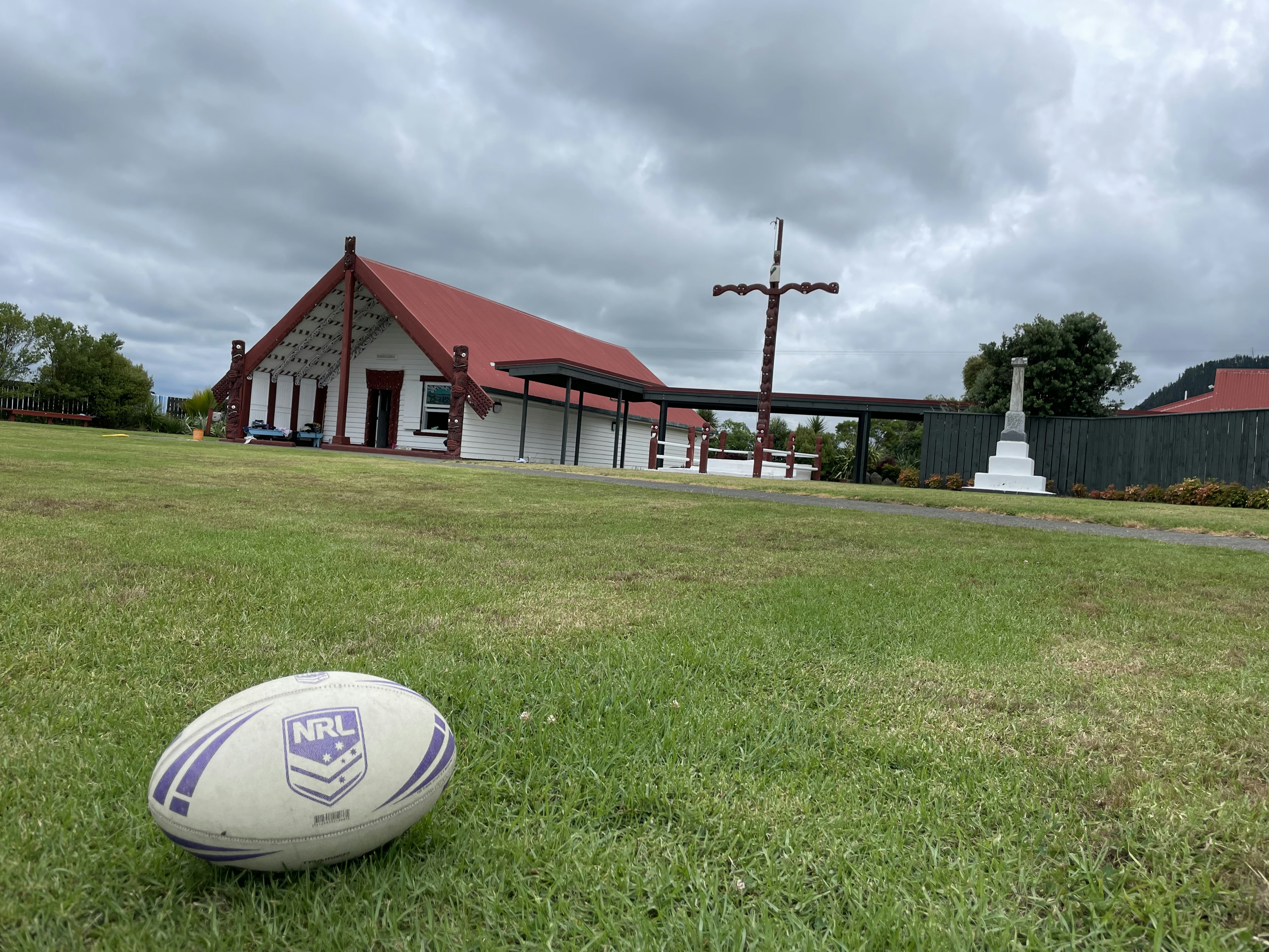 Sebuah bola rugby di atas rumput dengan bangunan atap merah dan langit mendung di latar belakang