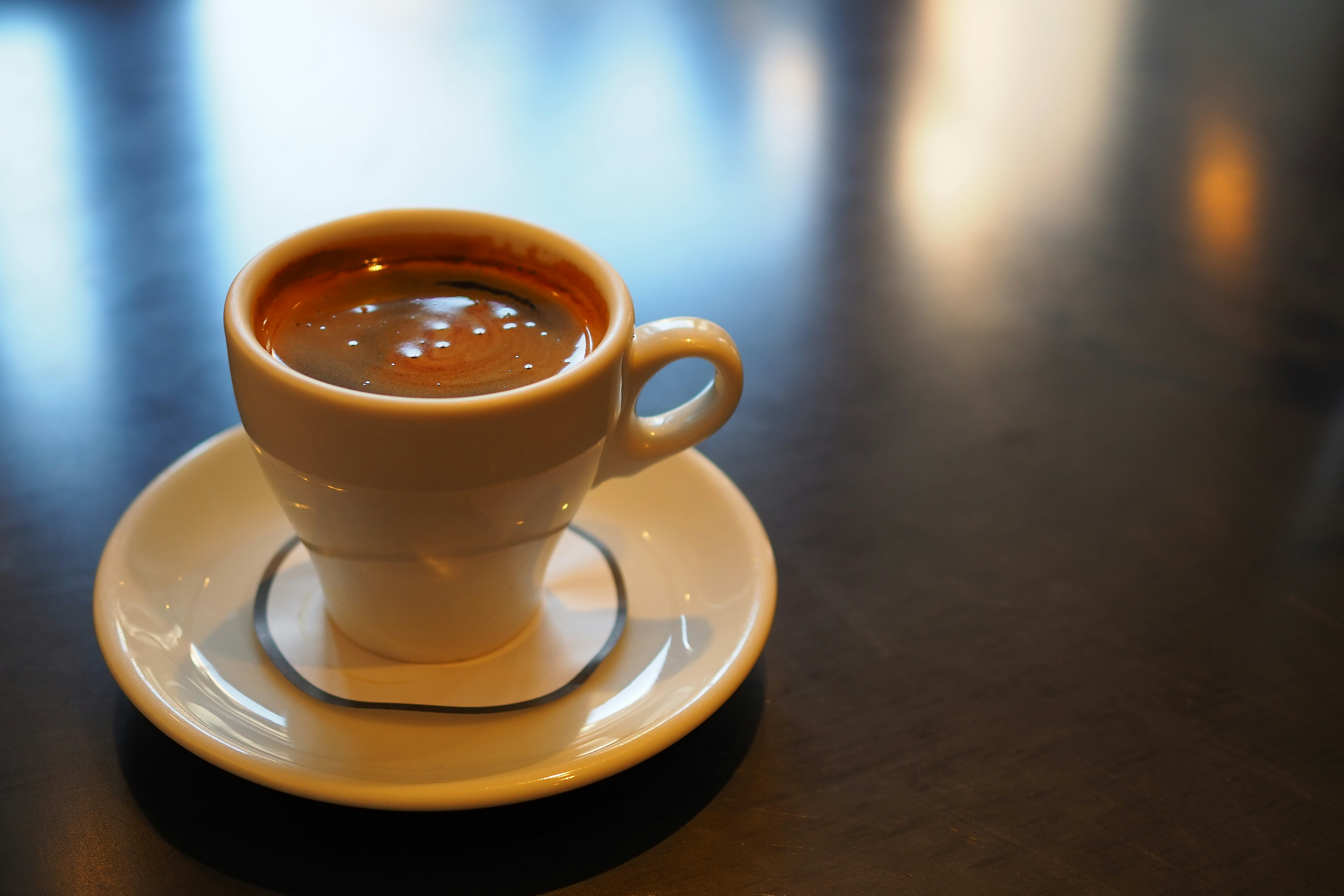 A white cup of coffee on a white saucer placed on a dark table