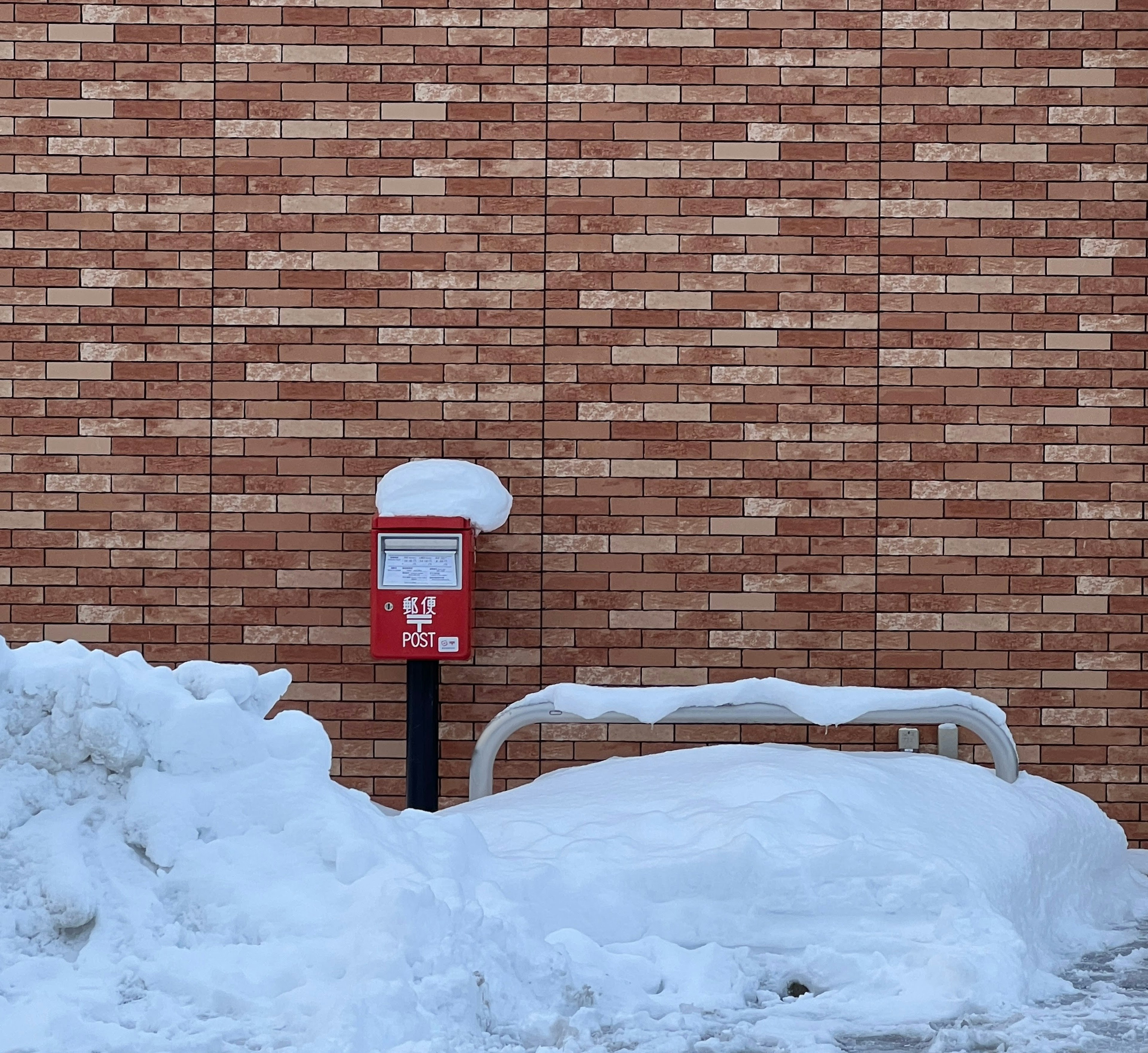 Red mailbox and snow-covered bench against a brick wall