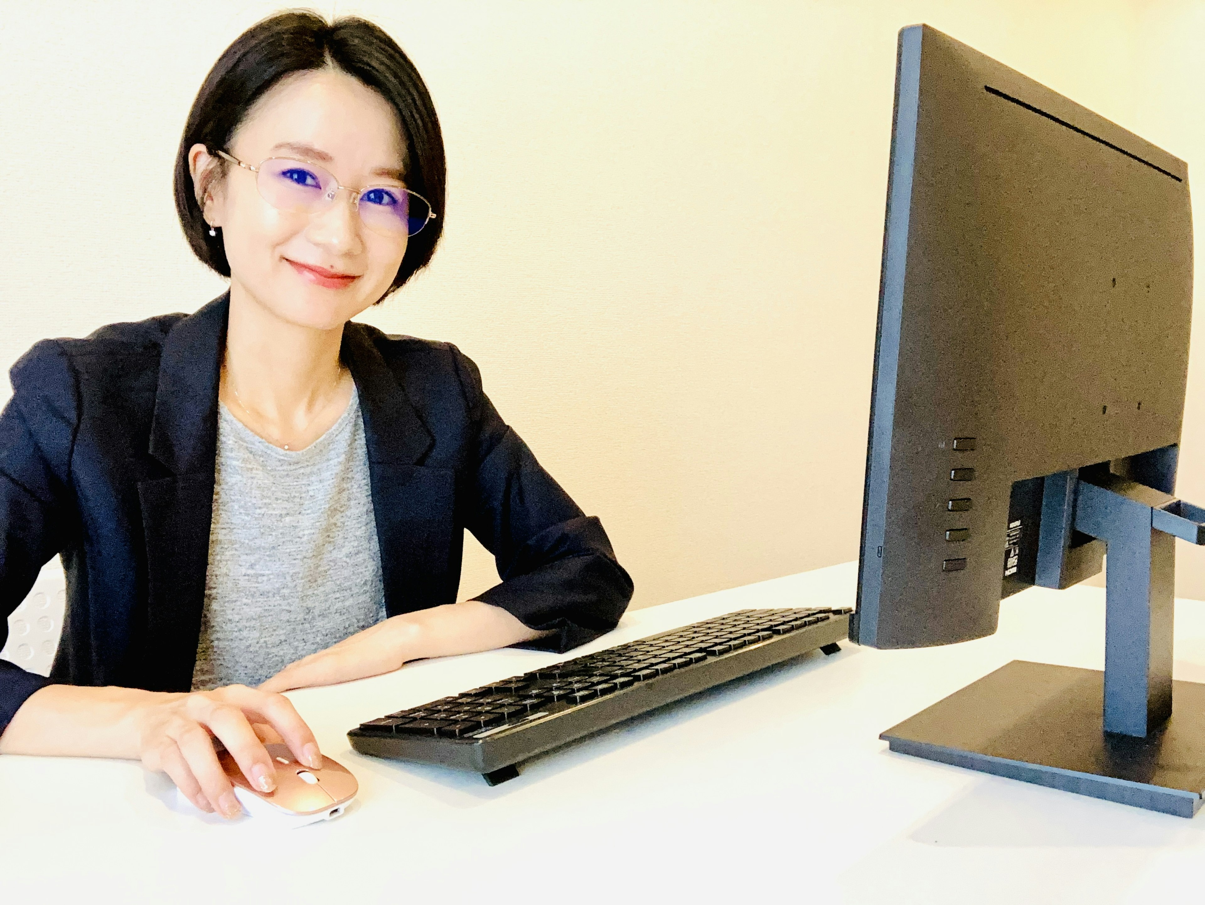 Una mujer sentada frente a una computadora con una camiseta gris y un blazer usando gafas