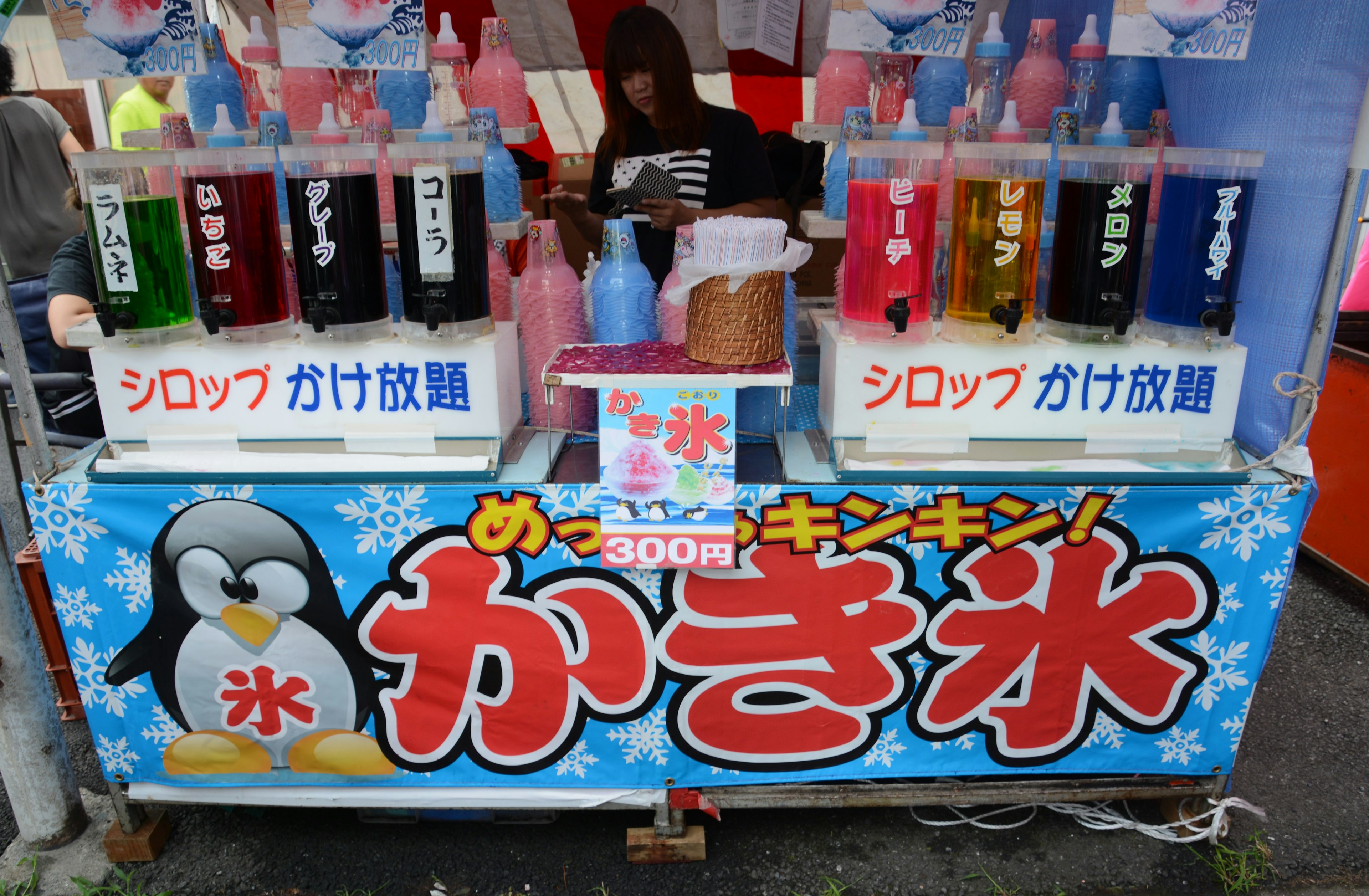 Bouteilles de sirop colorées exposées à un stand de kakigori avec une illustration de pingouin