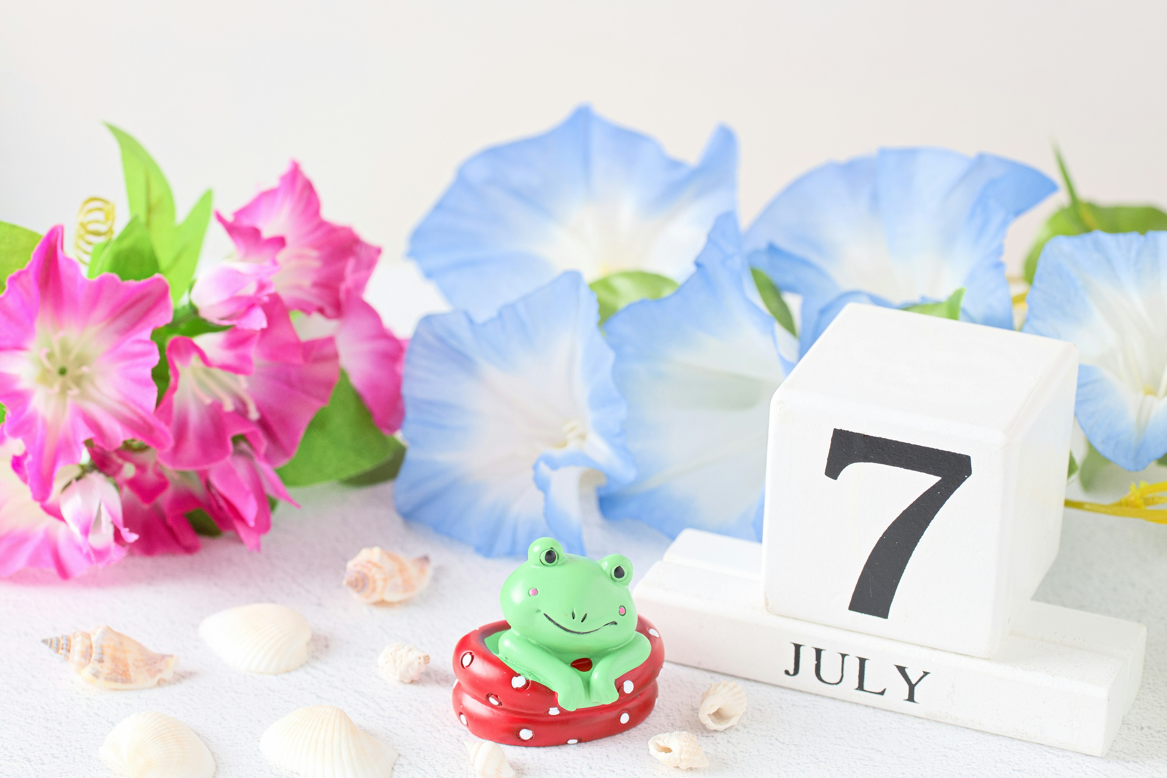 Une figurine de grenouille verte sur une base rouge avec un bloc de calendrier affichant le 7 juillet entouré de fleurs bleues et roses