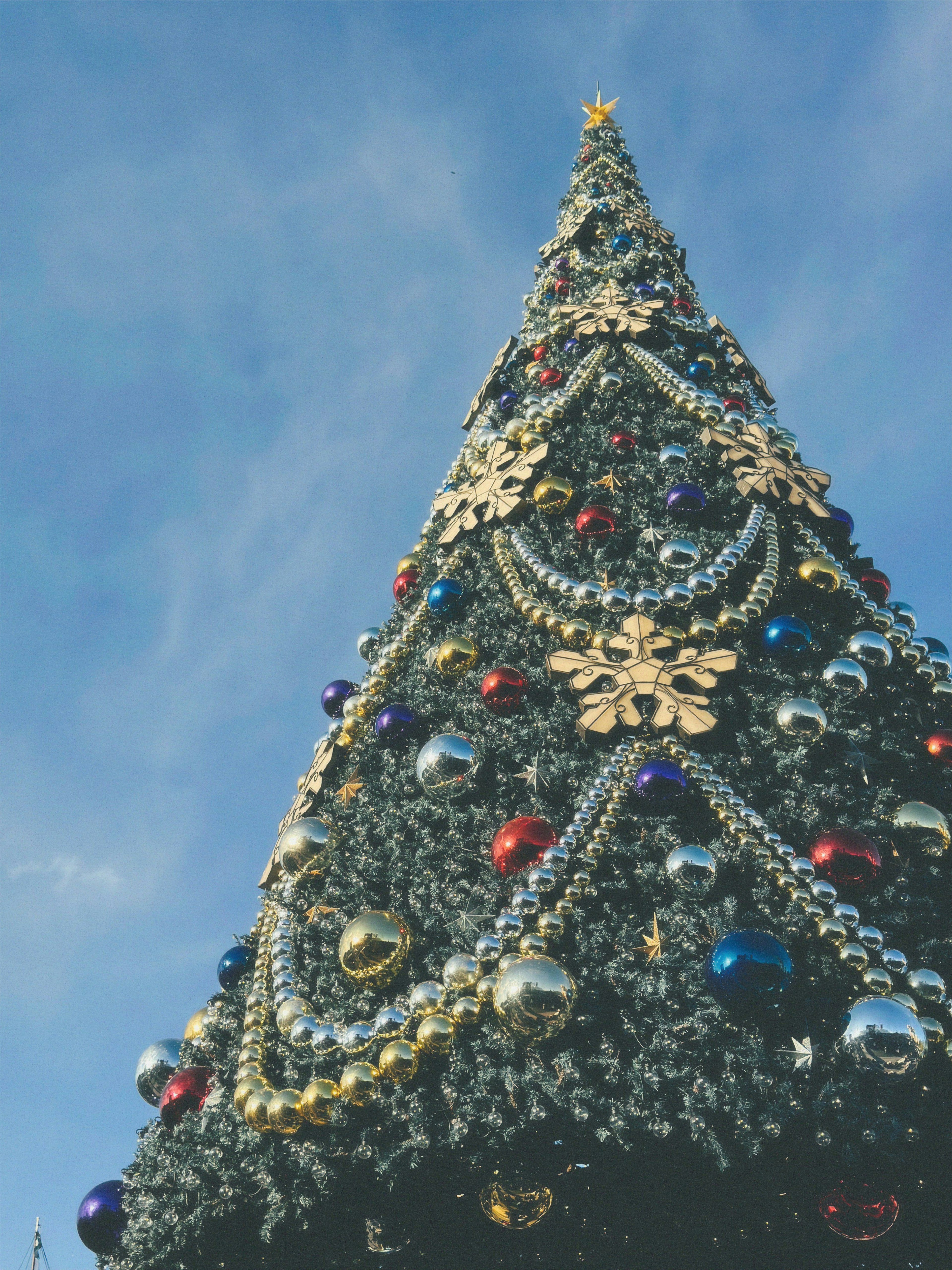 青空の下に立つ華やかなクリスマスツリー装飾には金色の雪の結晶とカラフルなオーナメントが飾られています