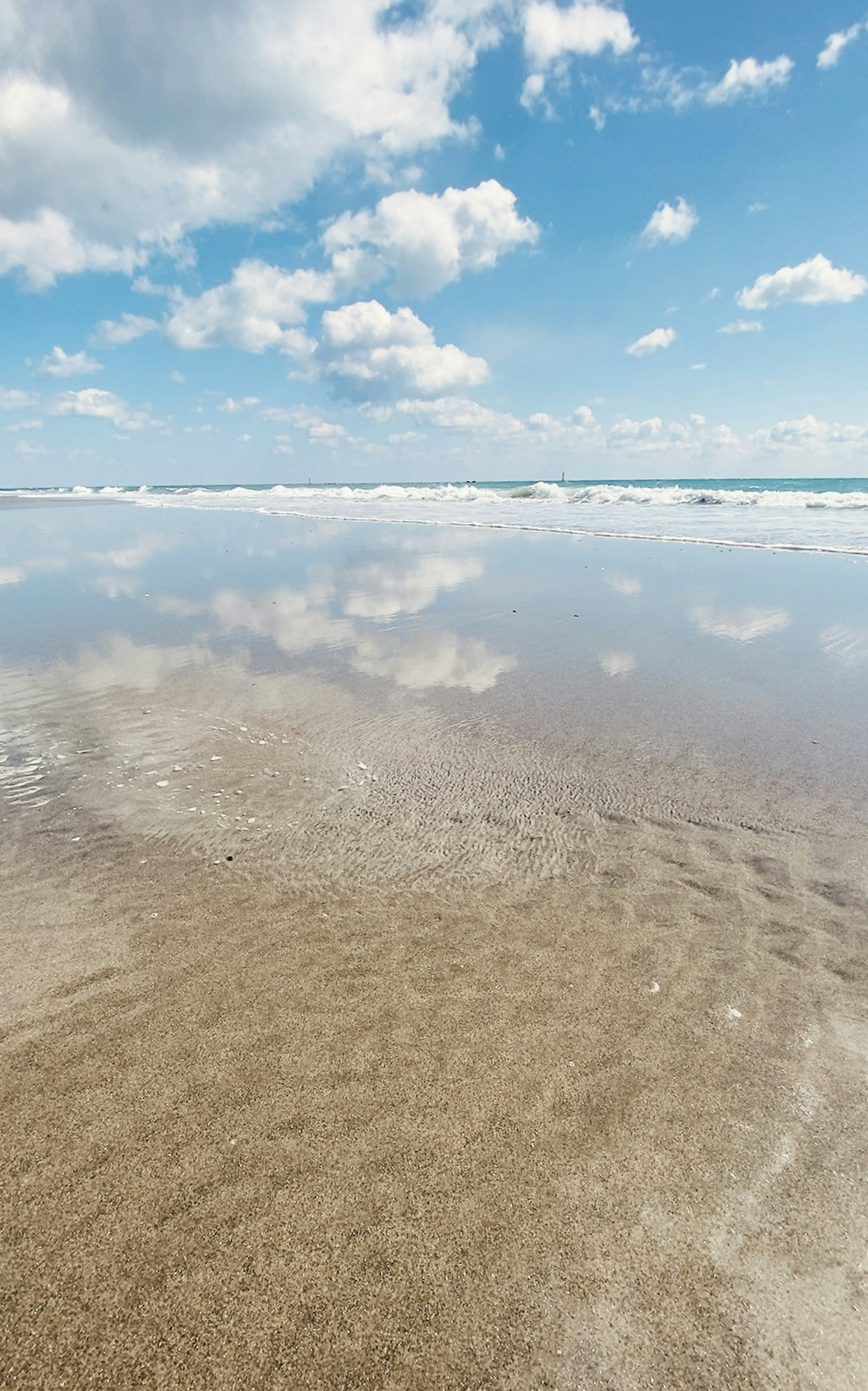 Pemandangan pantai tenang dengan refleksi awan putih di langit biru di atas air