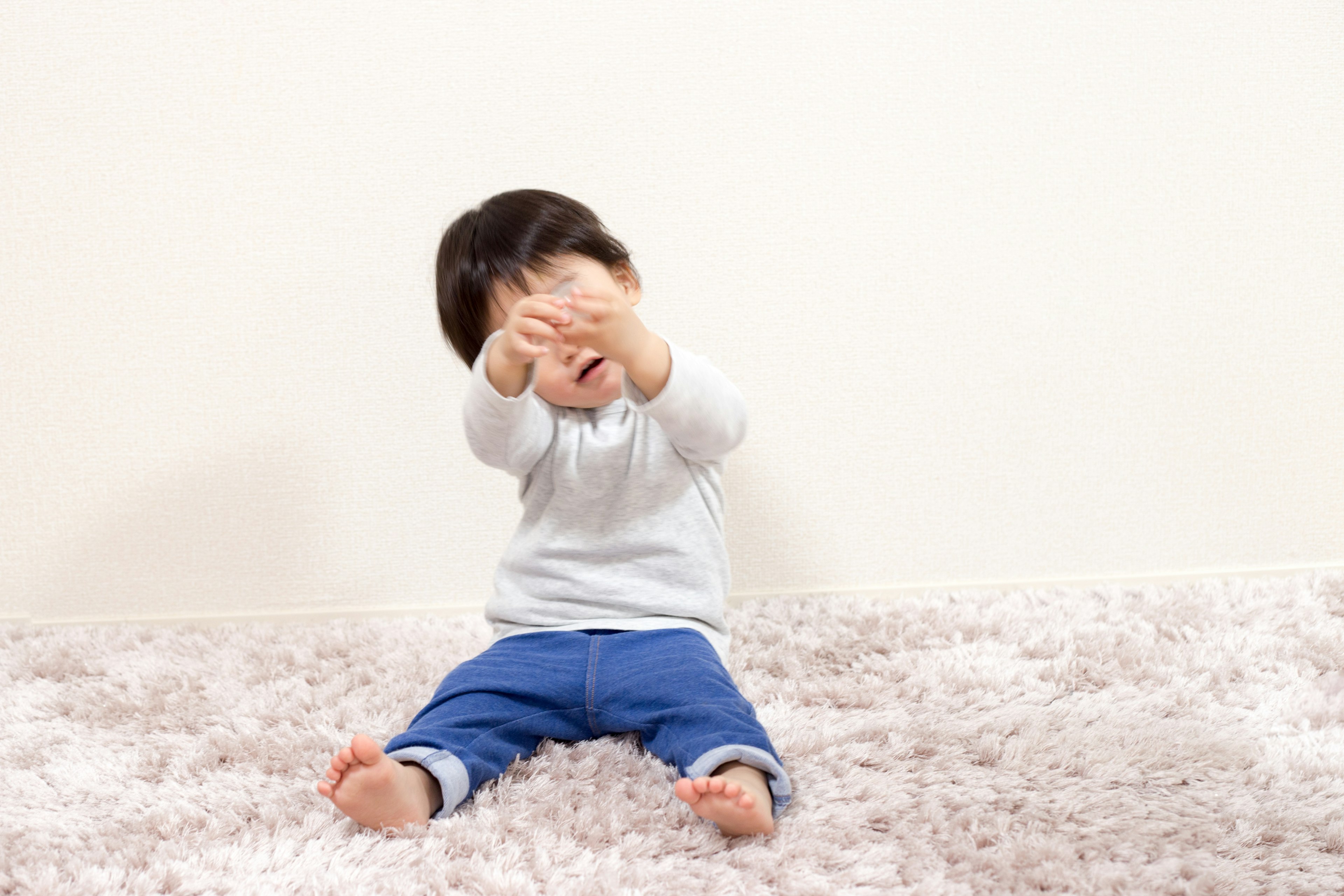 Seorang anak duduk di atas karpet lembut di depan dinding putih menutupi wajahnya dengan tangan