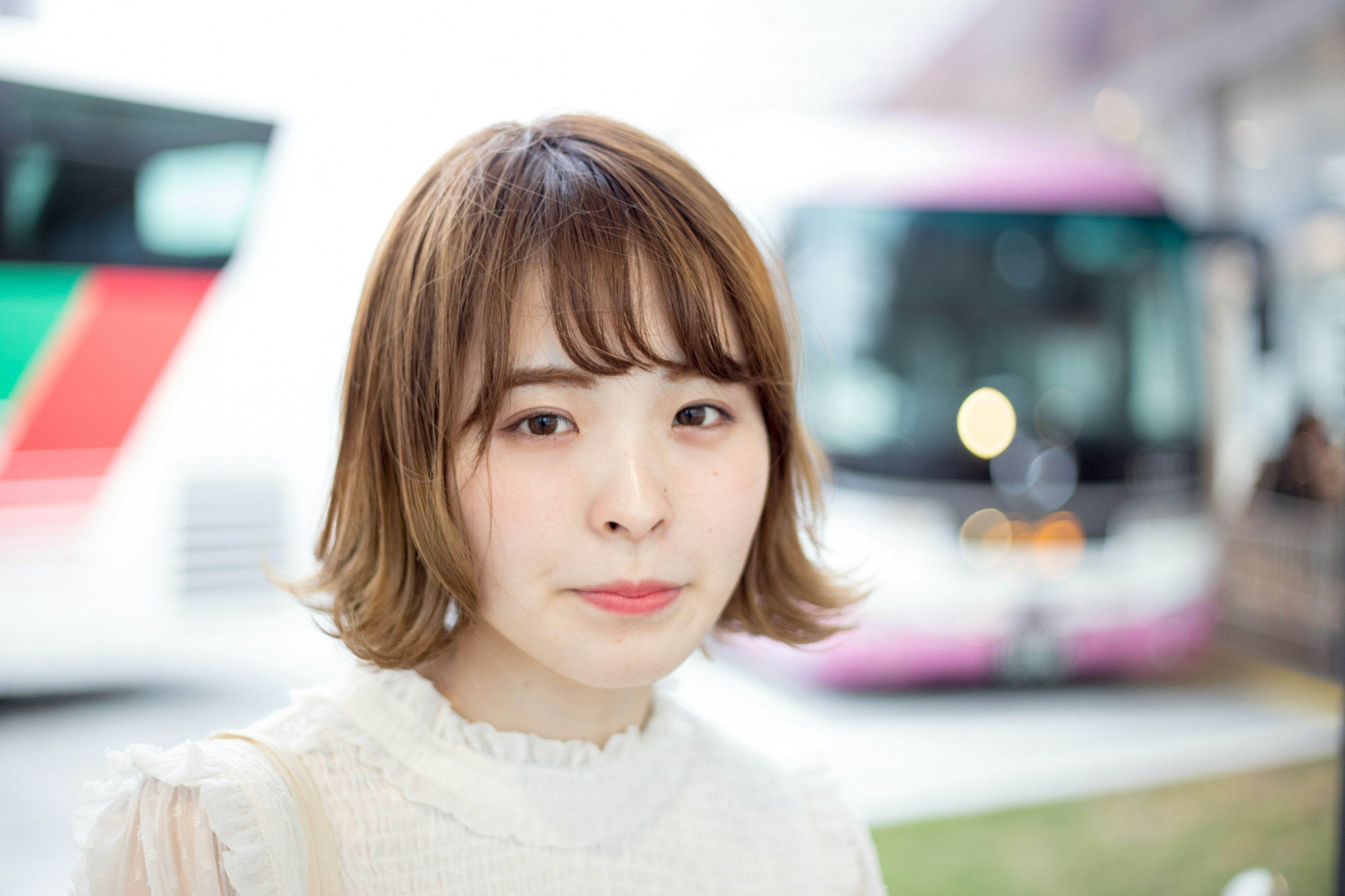 Eine Frau mit kurzen braunen Haaren in einem weißen Outfit lächelt mit einem Bus im Hintergrund