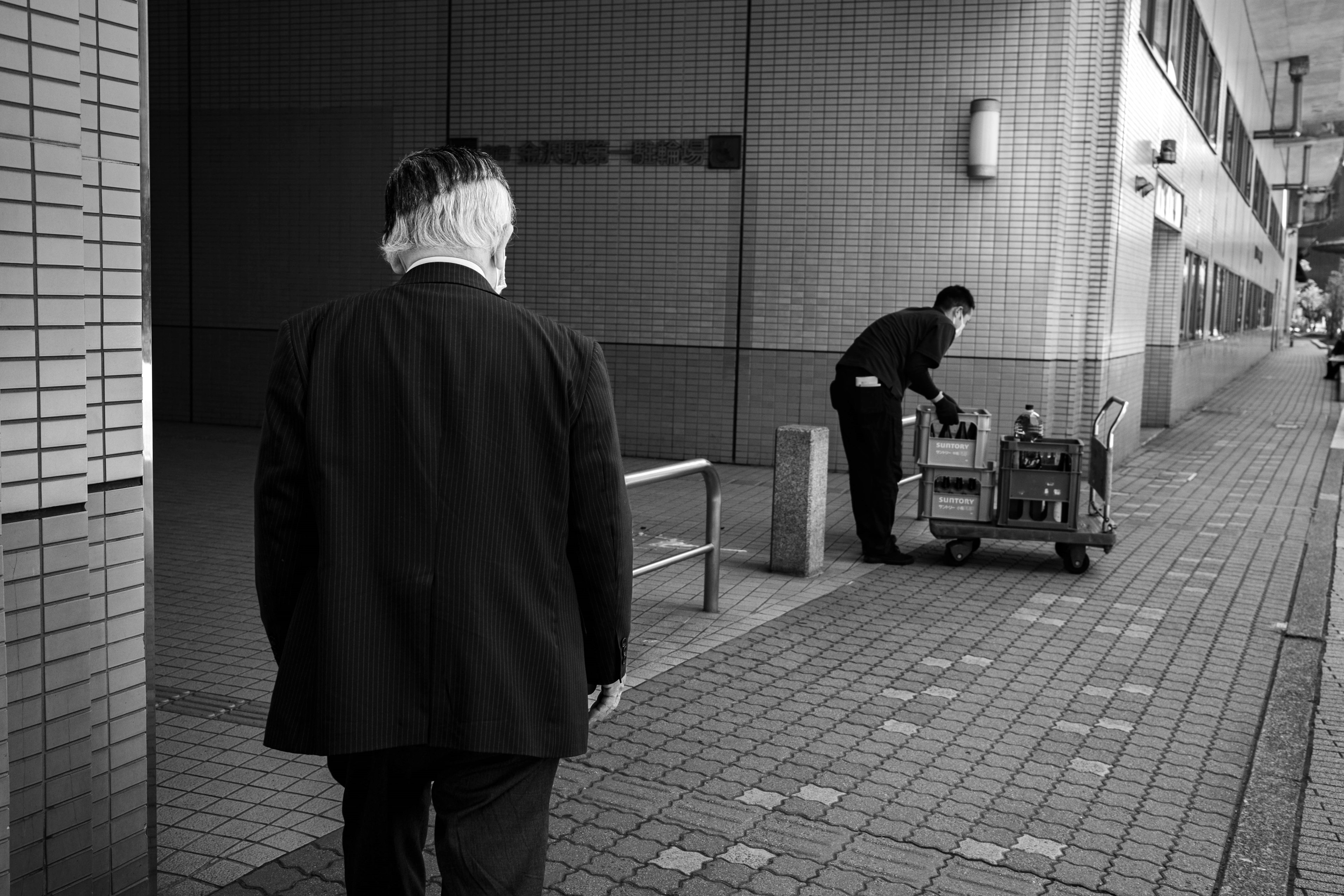 A man in a suit walking away in a black and white urban setting with another man working nearby