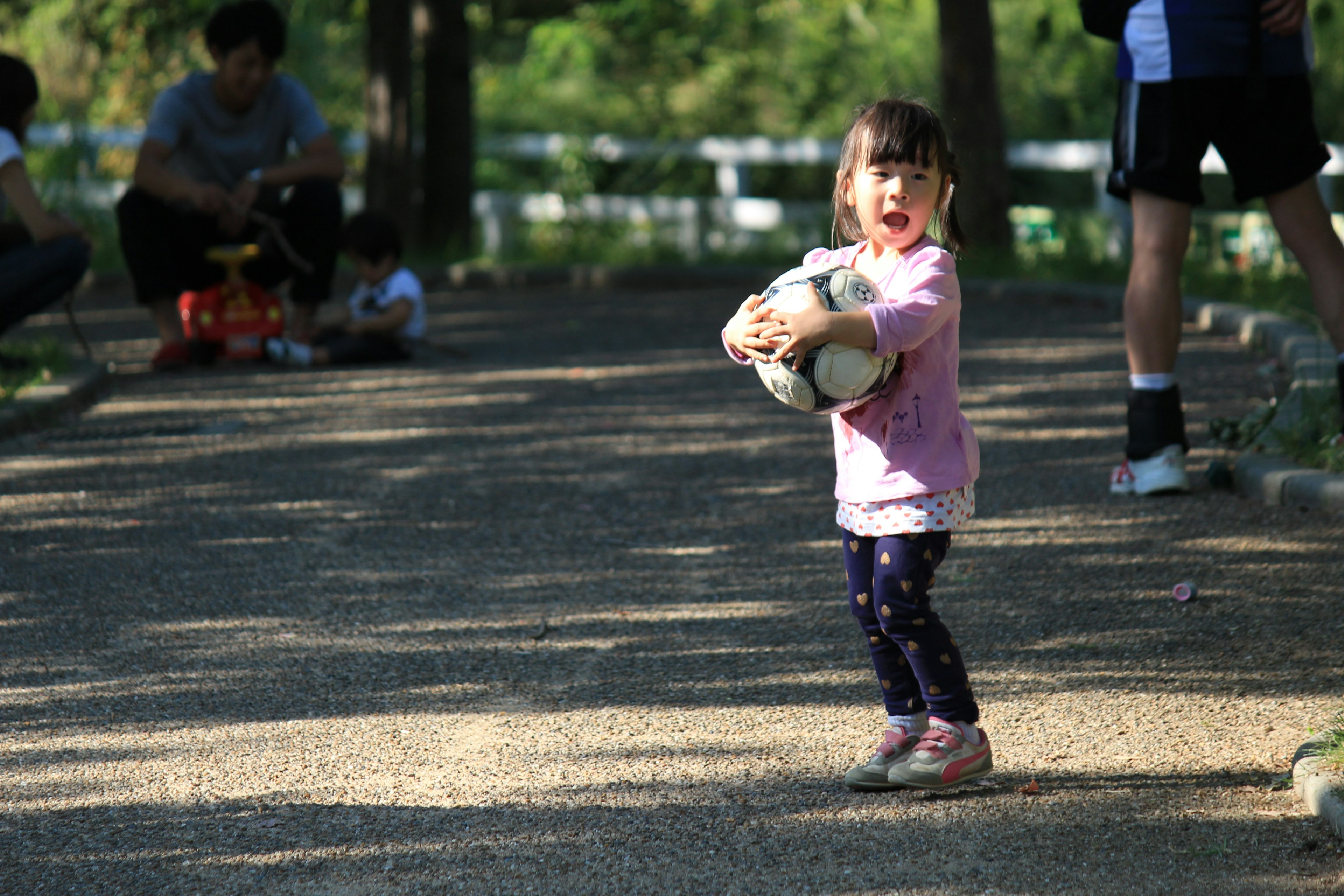 Young girl holding a soccer ball in a park surrounded by people