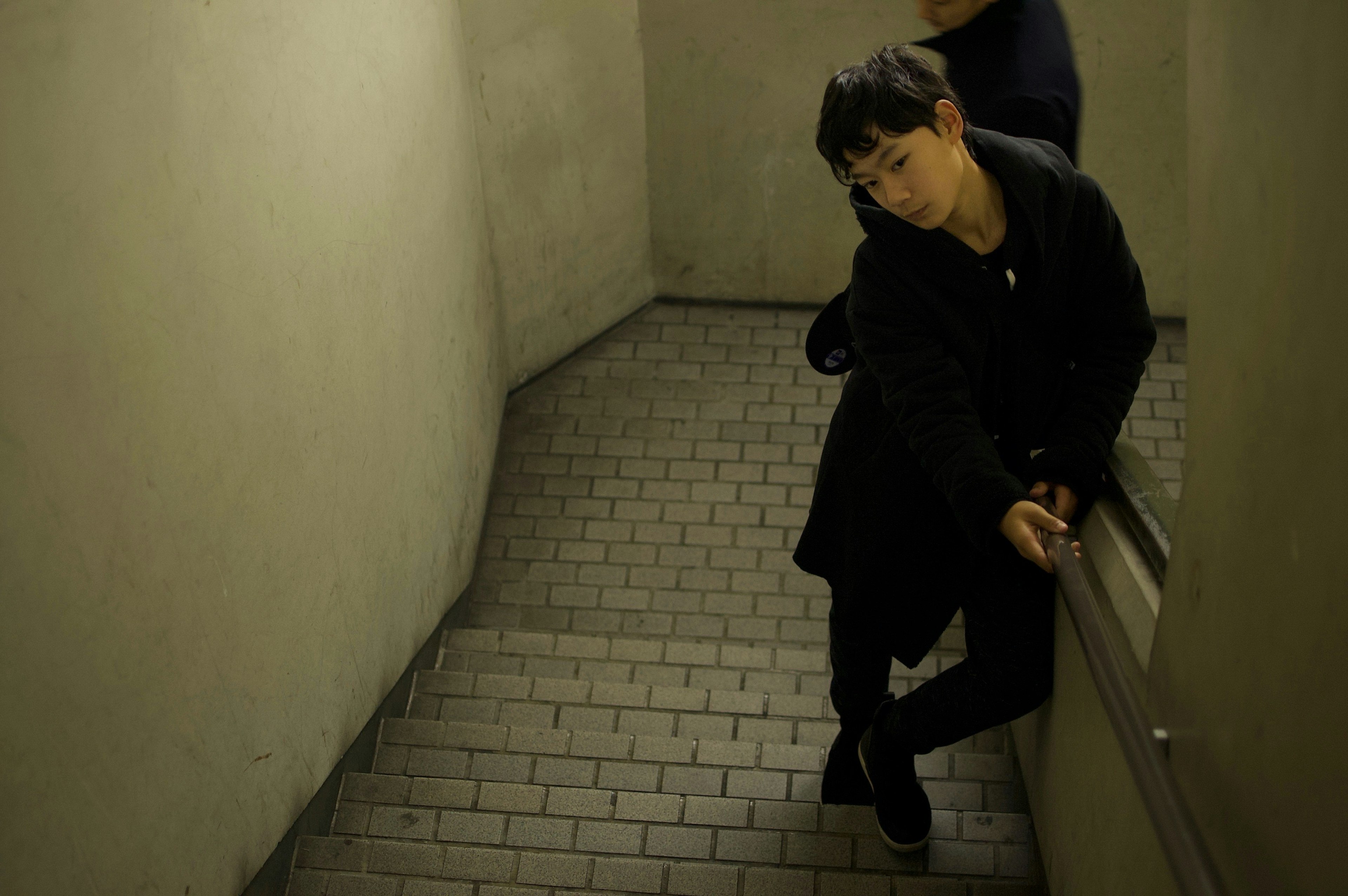 Man leaning on a staircase wearing a black coat in dim lighting