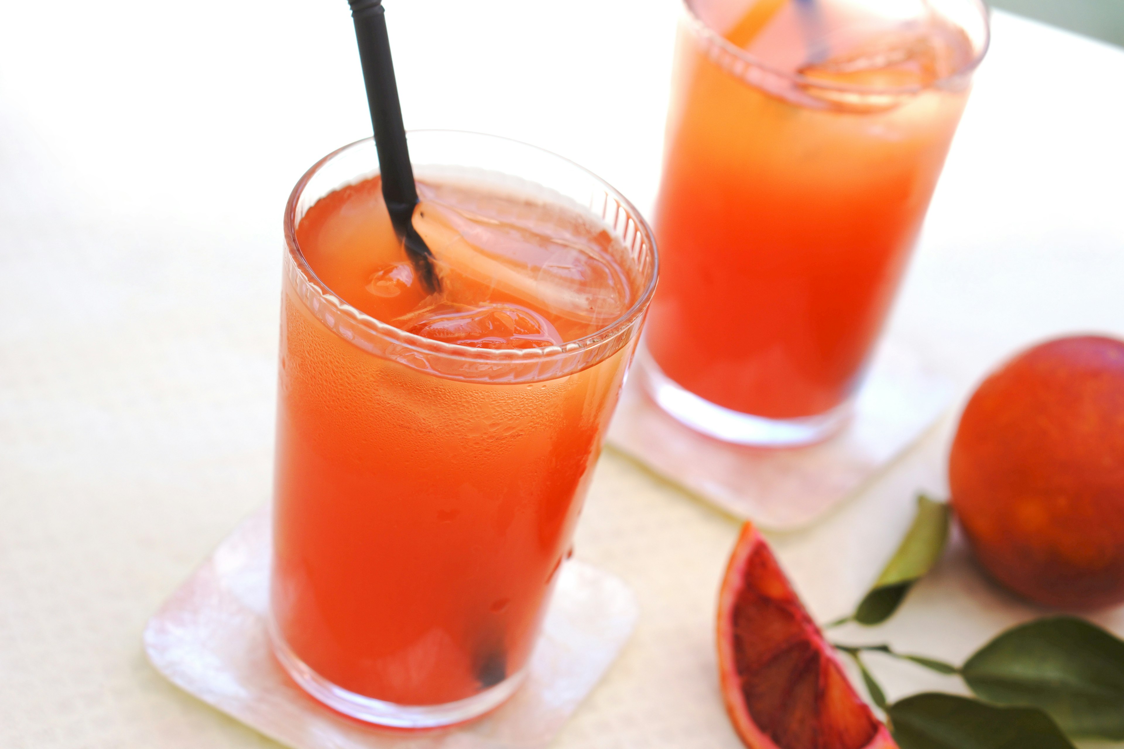 Two glasses of vibrant orange cocktail on a table with a black straw and blood orange slice