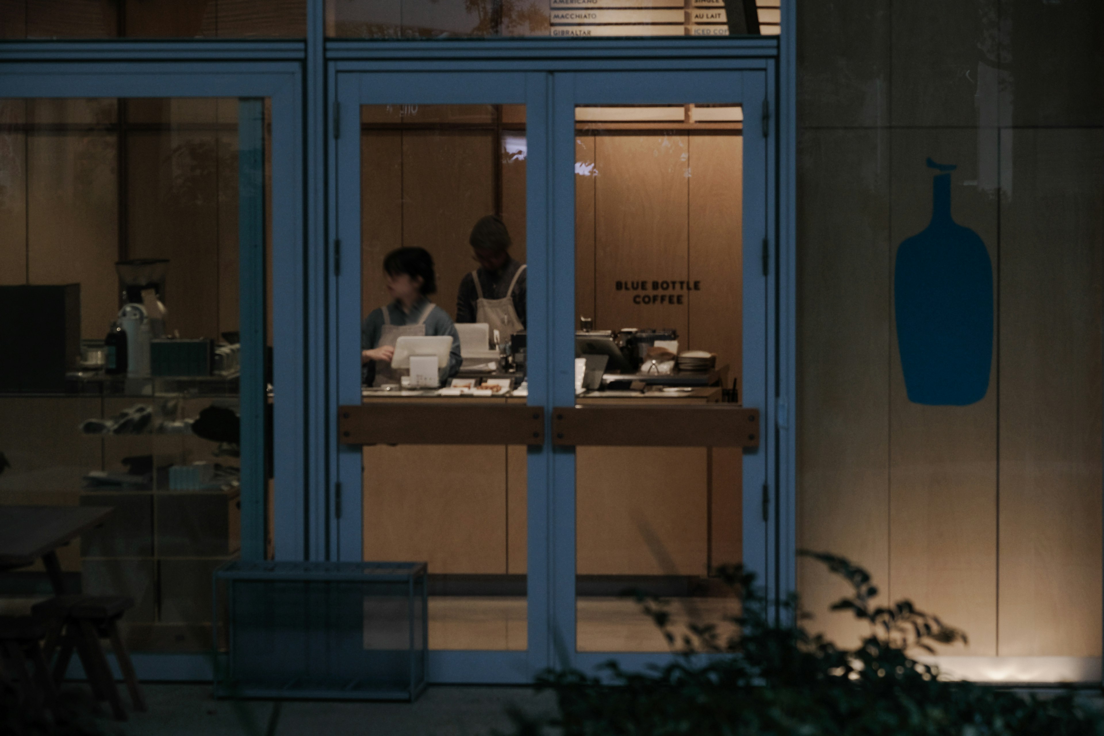 Cafe interior viewed through a window with staff working and simple wooden decor