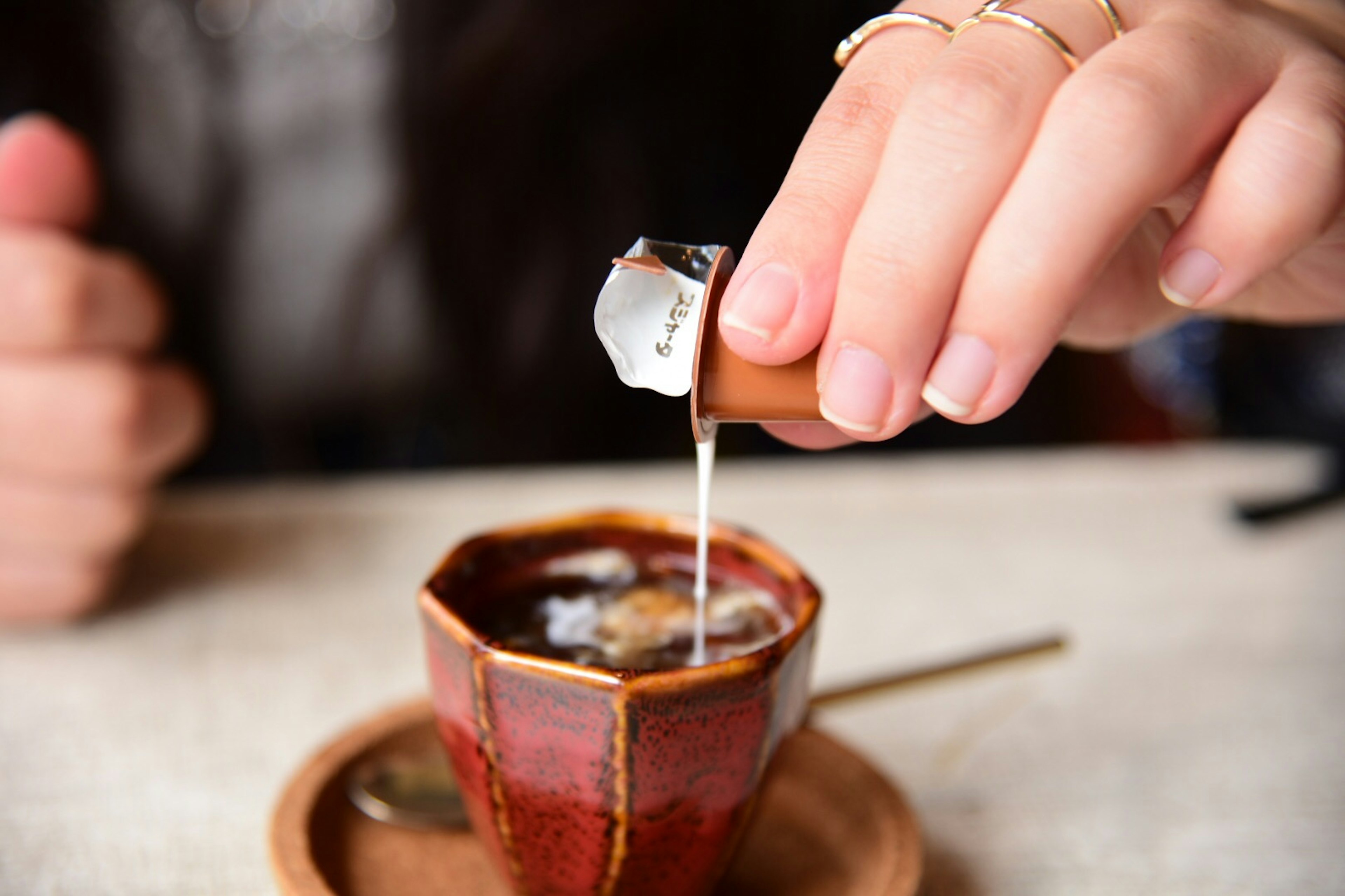 A hand pouring cream into a coffee cup with a red ceramic design