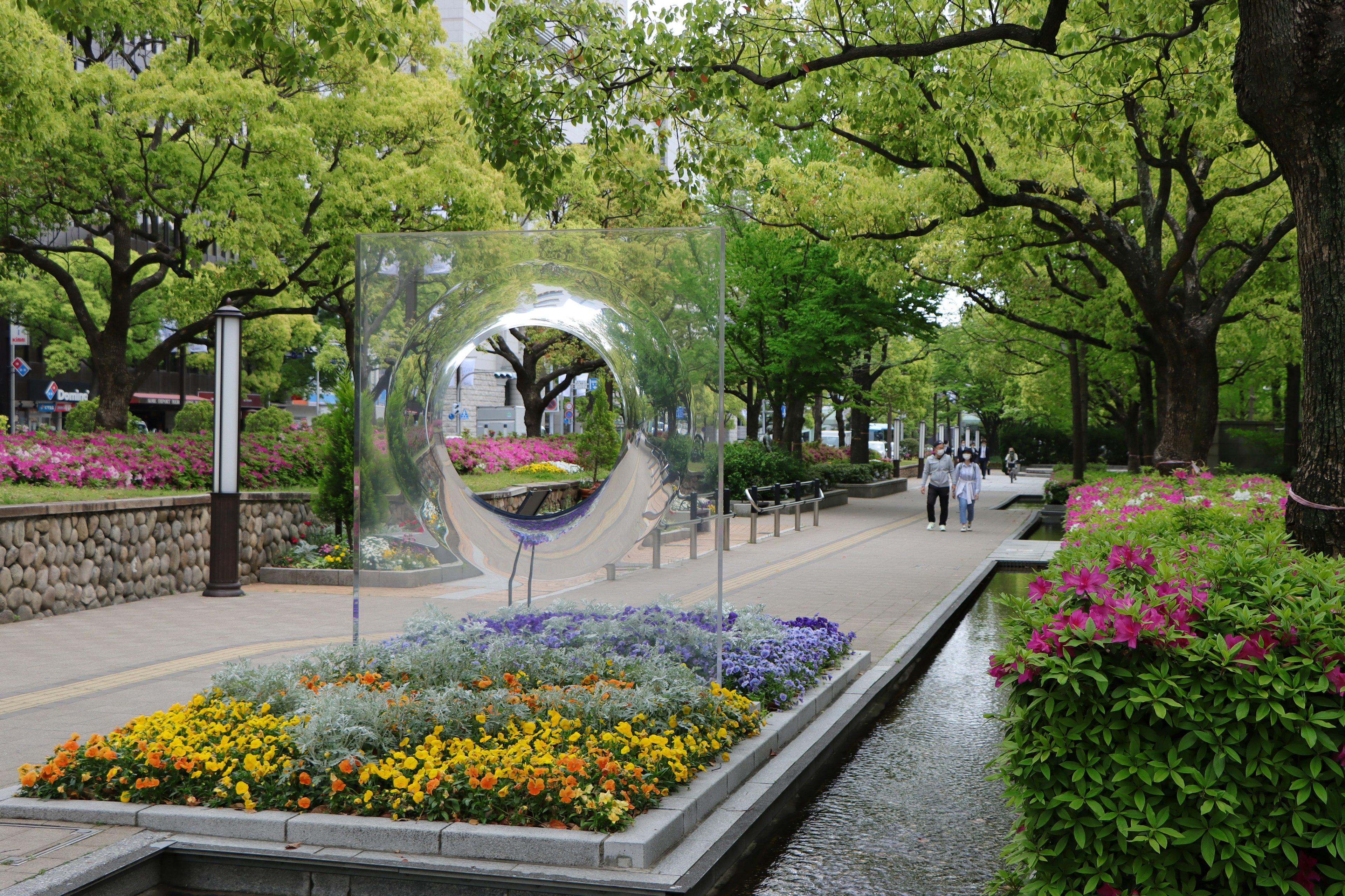 Vista escénica de un parque con vibrantes parterres de flores y árboles verdes exuberantes con una escultura transparente central