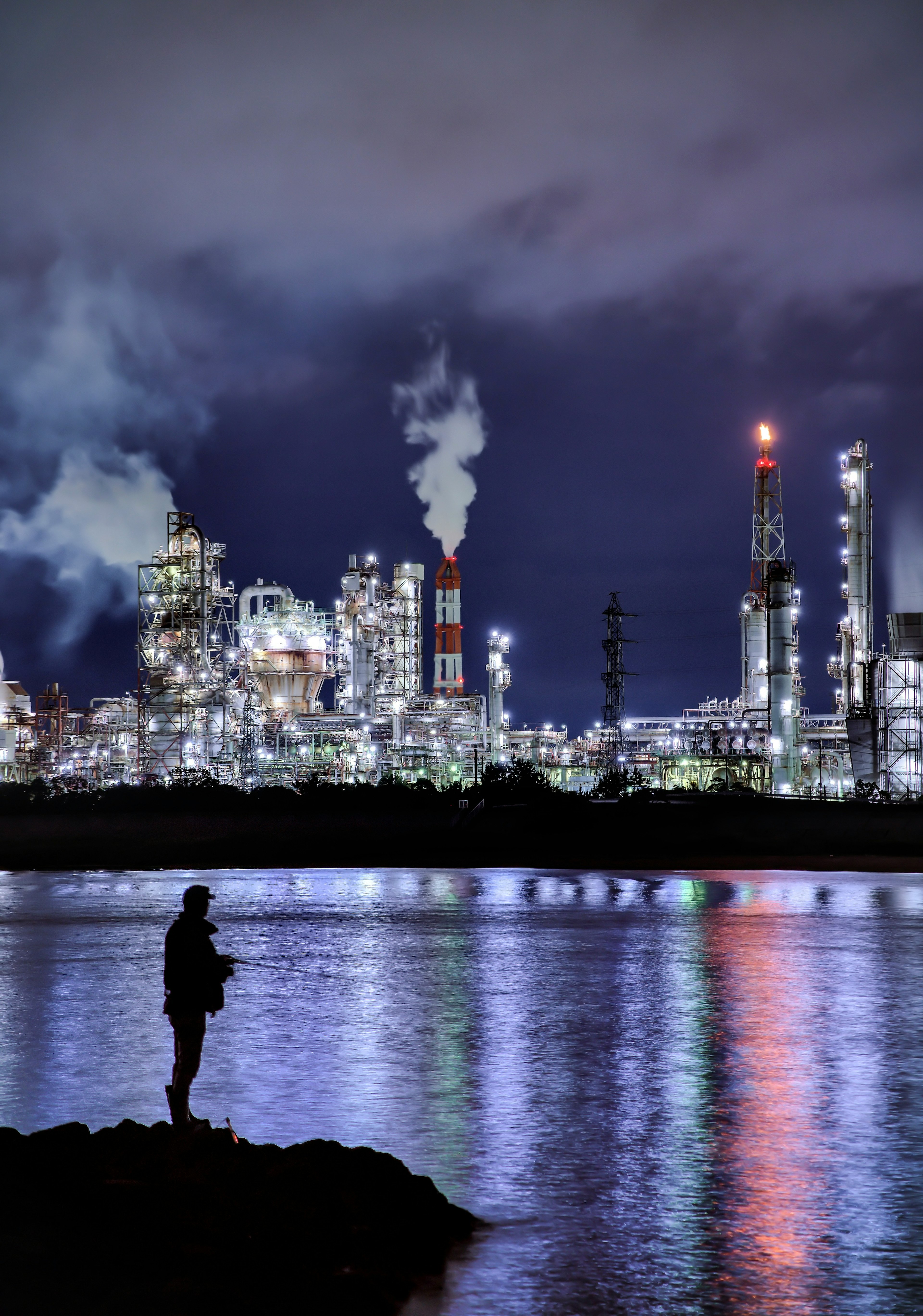 Silueta de un pescador junto al agua con un paisaje industrial de noche