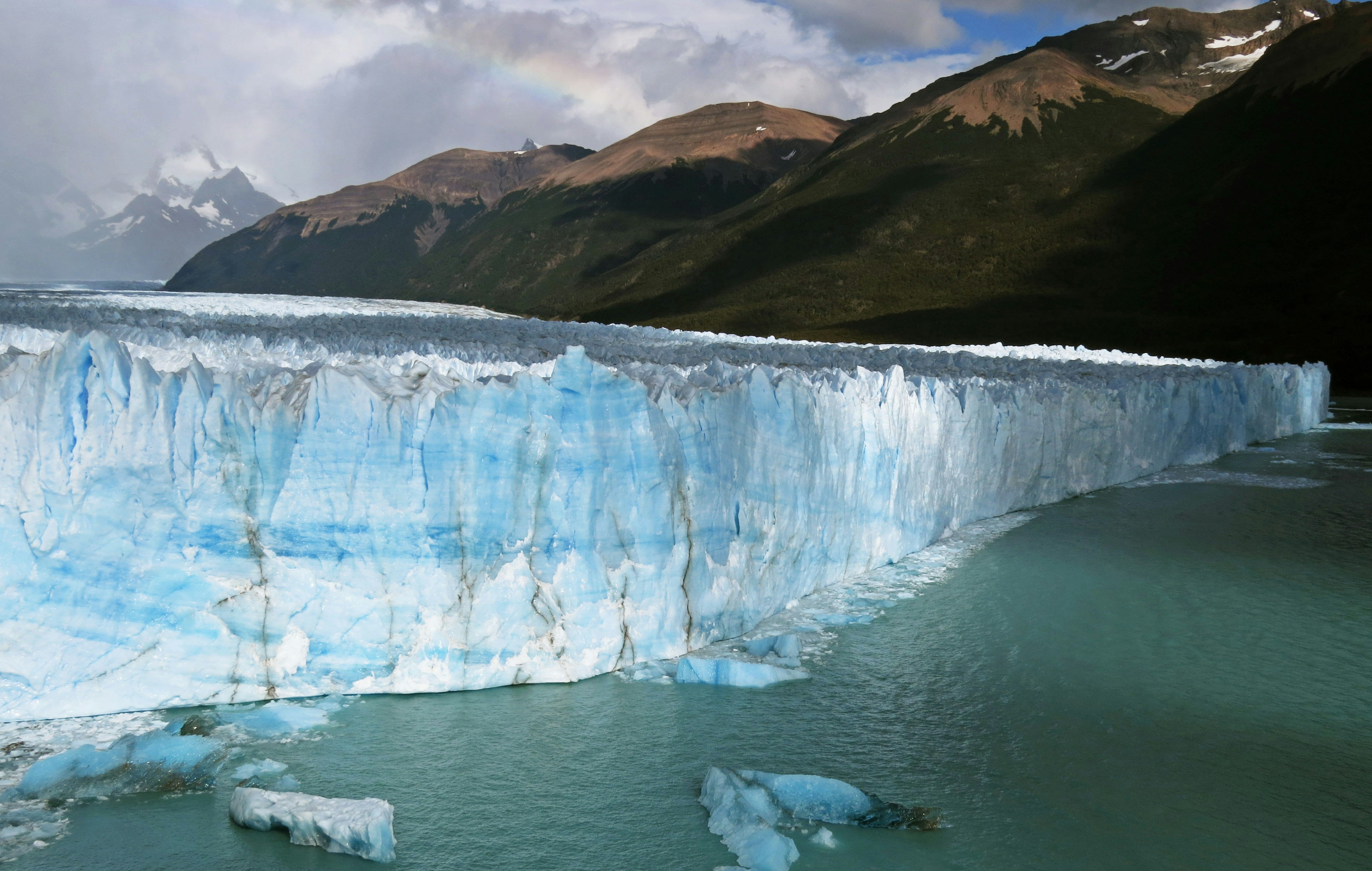 Pared de hielo azul de un glaciar con agua tranquila y montañas al fondo