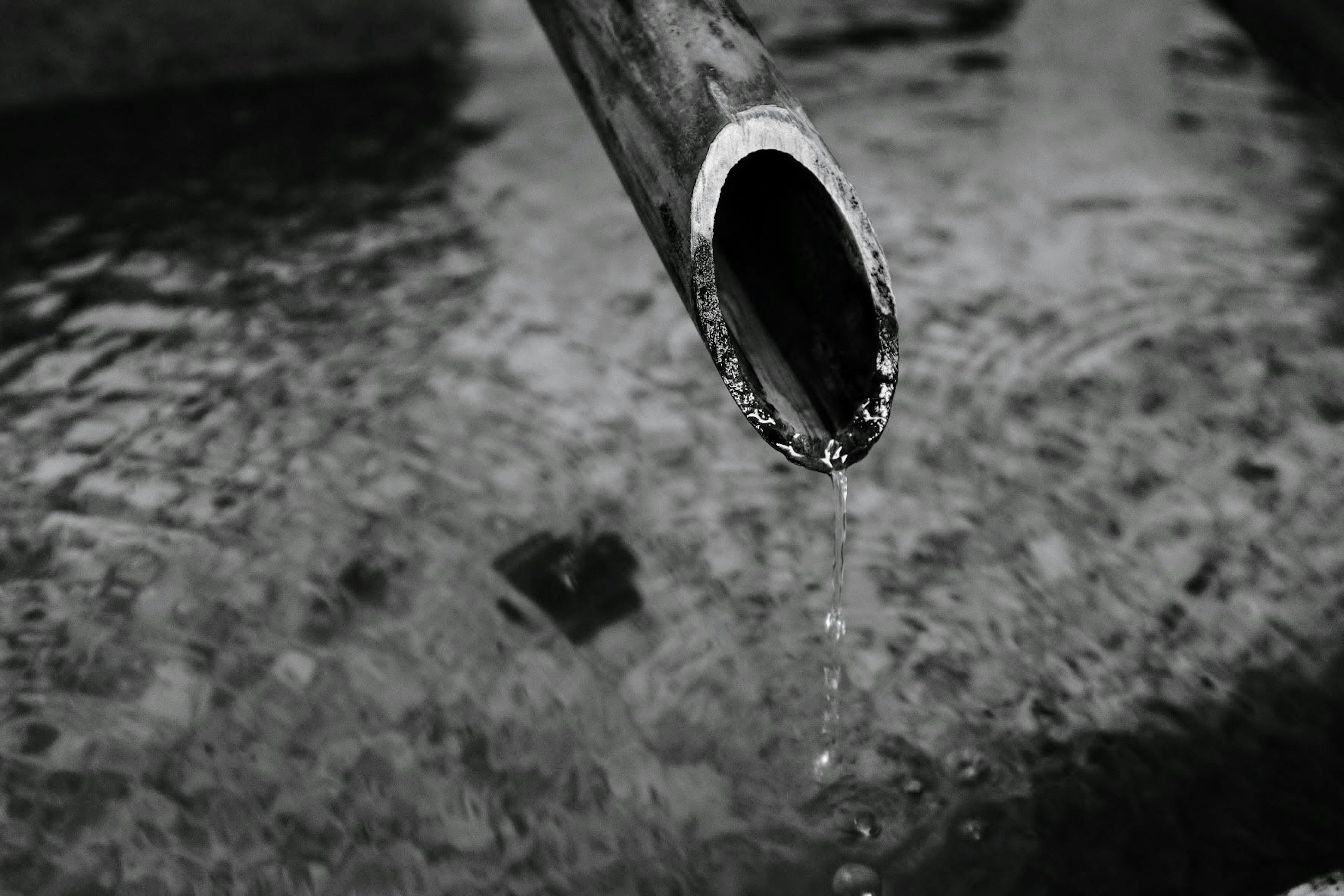 Nahaufnahme eines Metallrohrs, das Wasser auf eine wellende Oberfläche in Schwarzweiß tropft