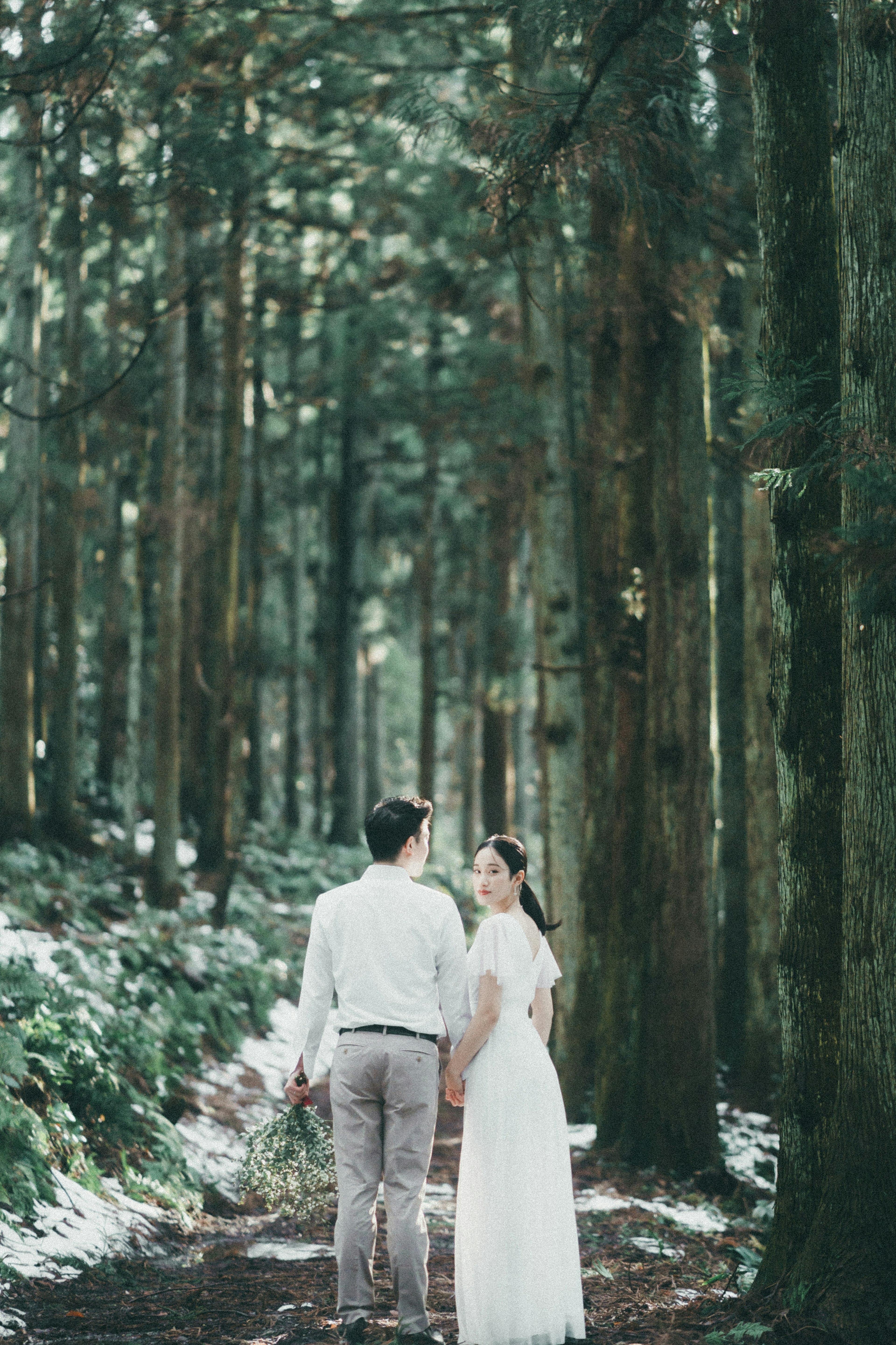 Couple in wedding attire standing in a lush forest looking back