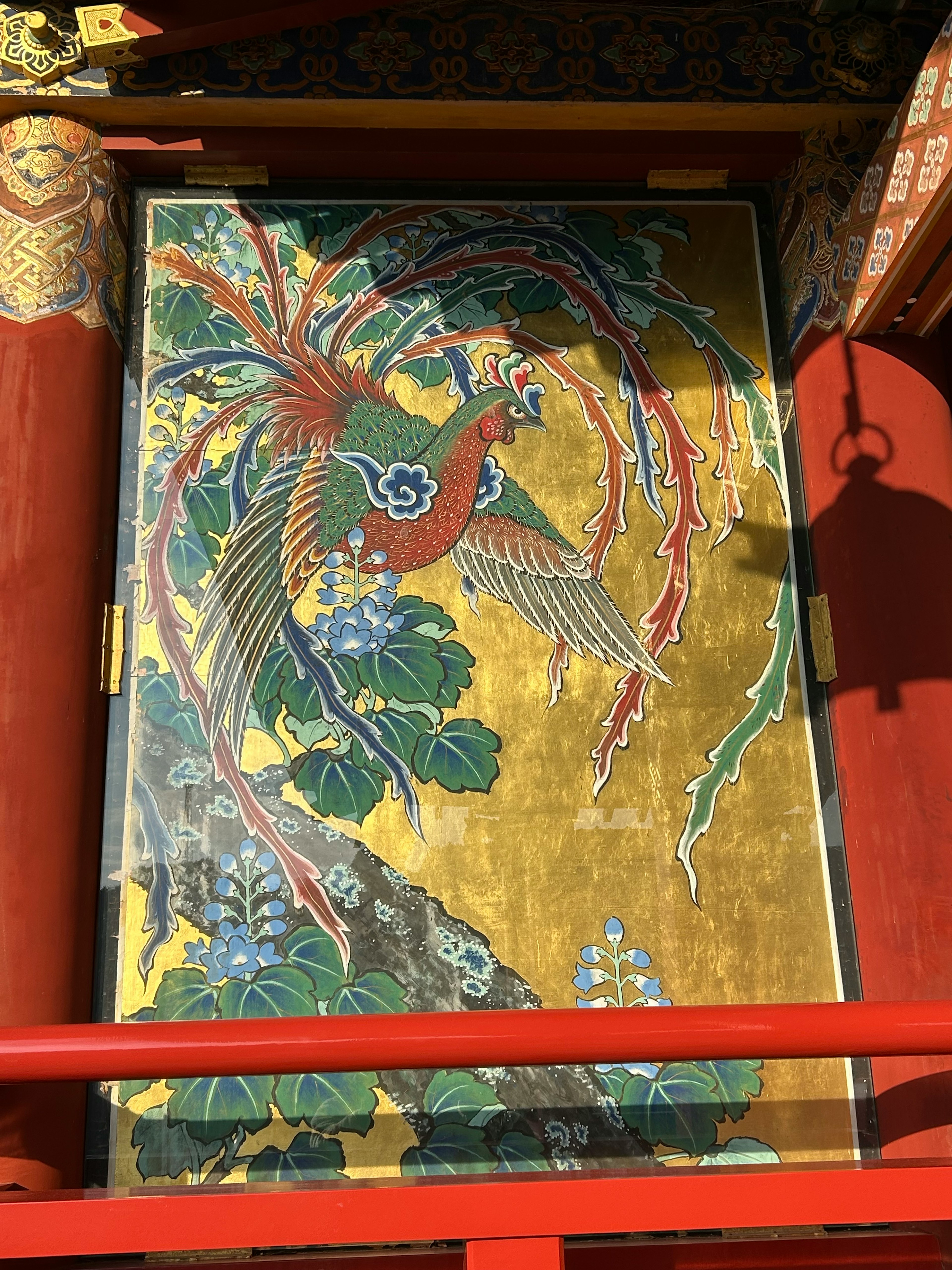 A vibrant phoenix painting with a golden background surrounded by green leaves and blue flowers