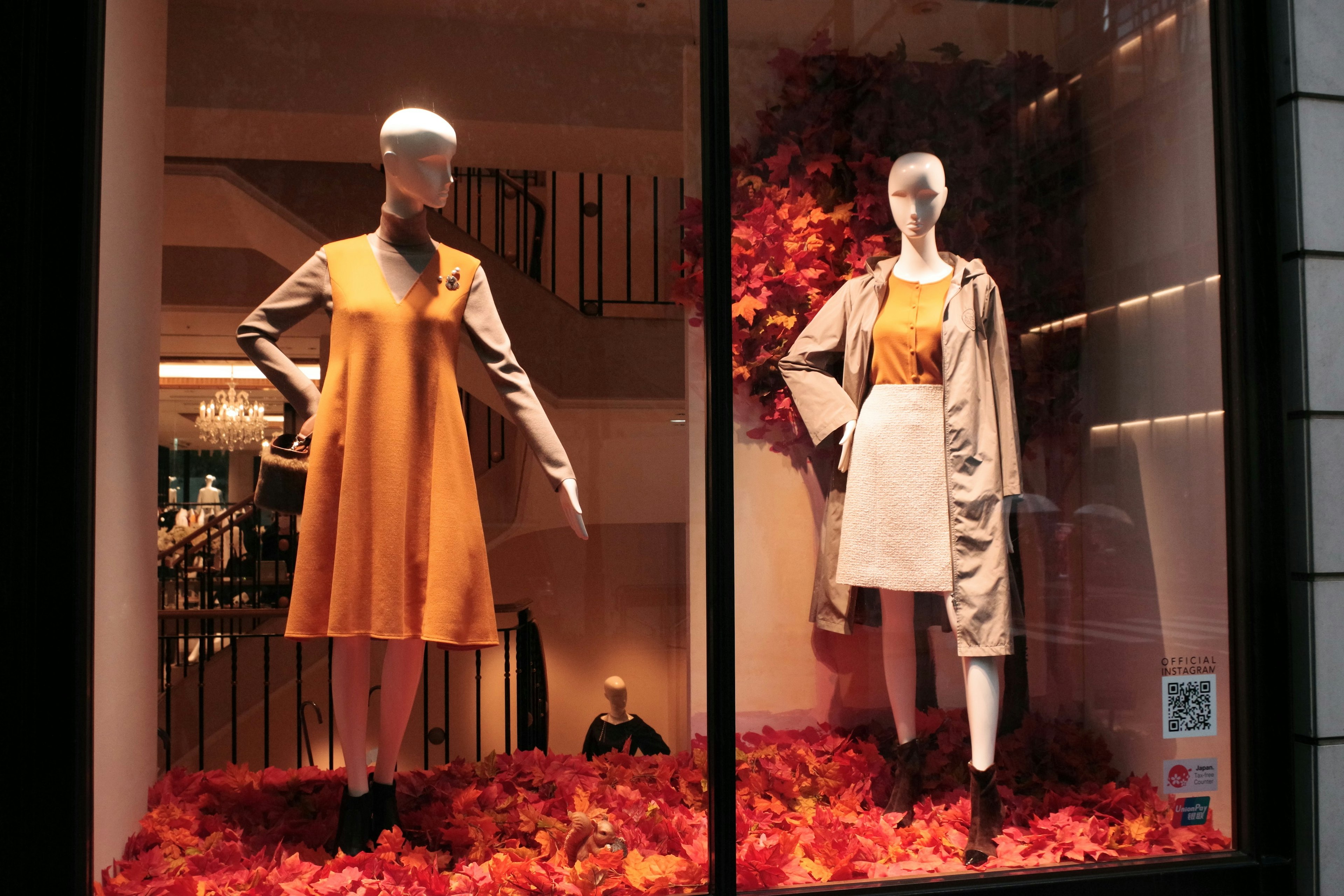 Zwei Schaufensterpuppen in einem Schaufenster tragen ein oranges Kleid und ein weißes Kleid umgeben von Herbstblättern