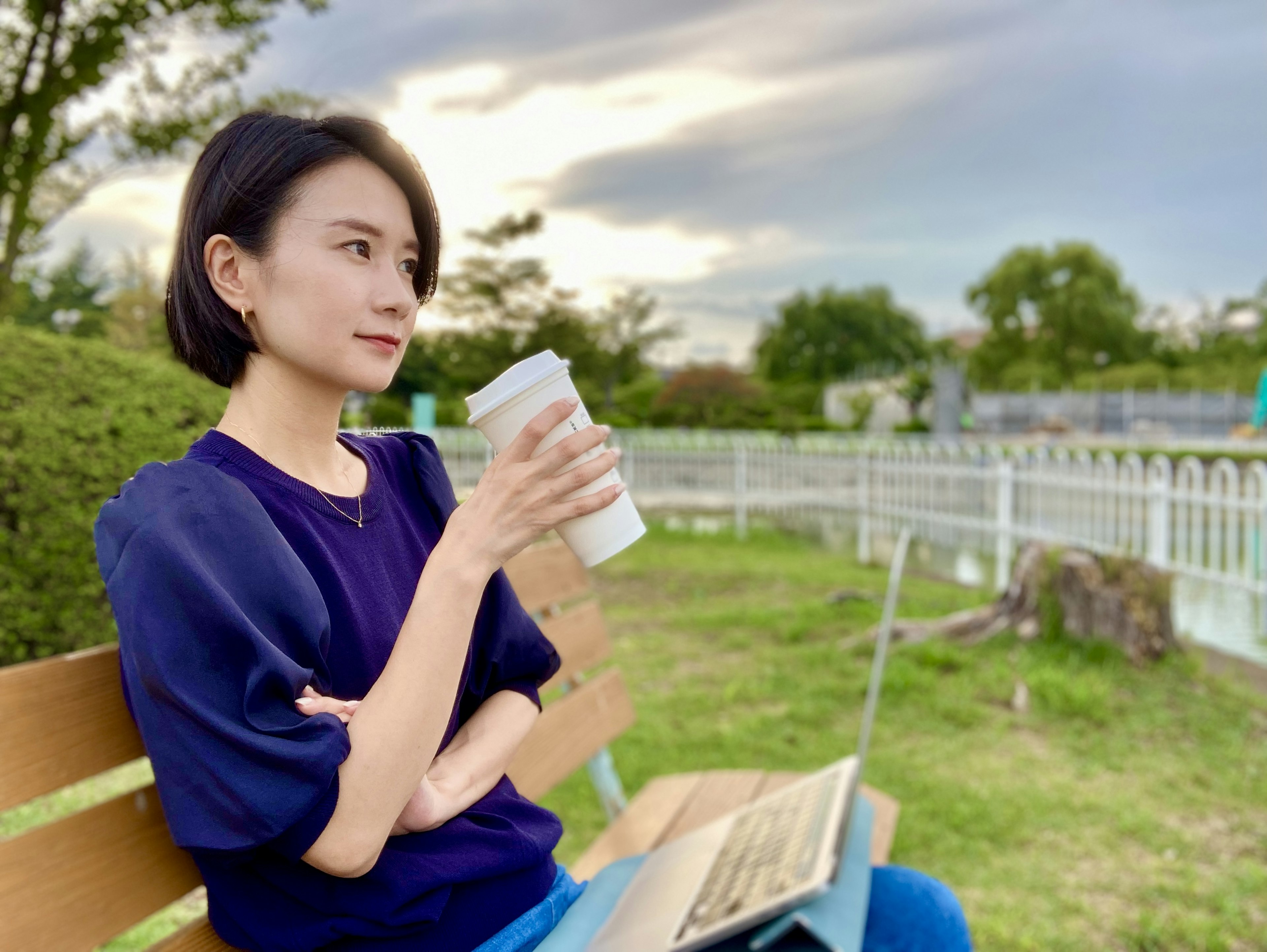 Woman sitting on a park bench holding coffee and using a laptop