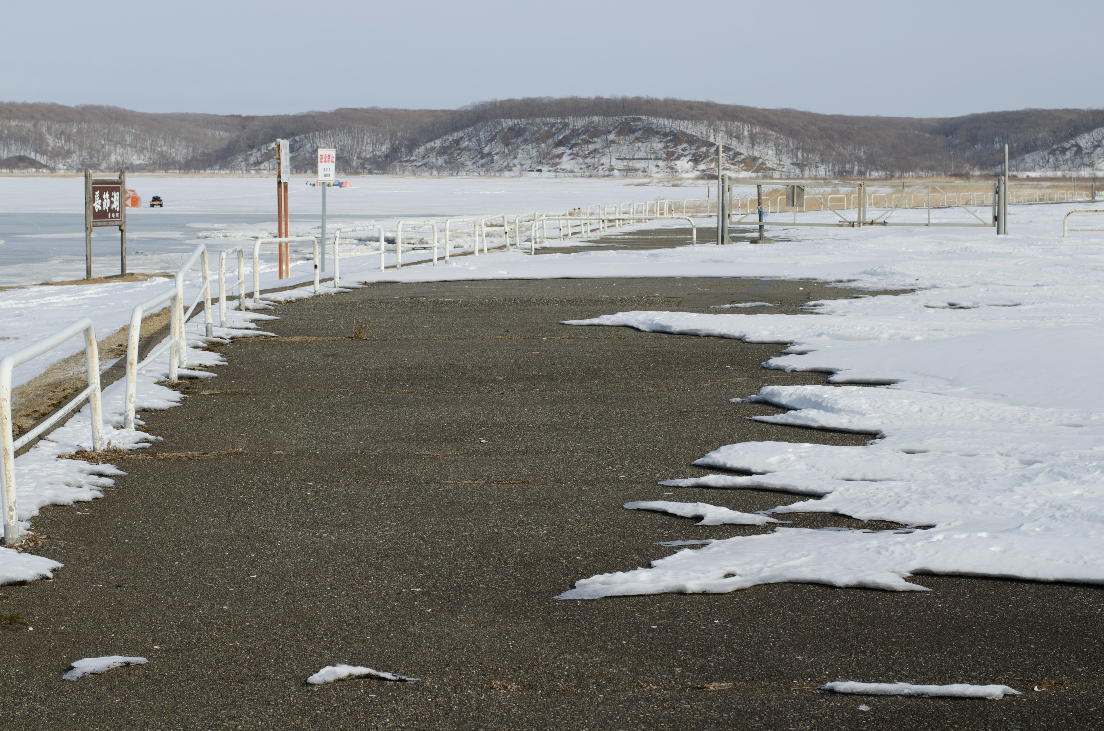 Paved path along a snow-covered shore with distant hills