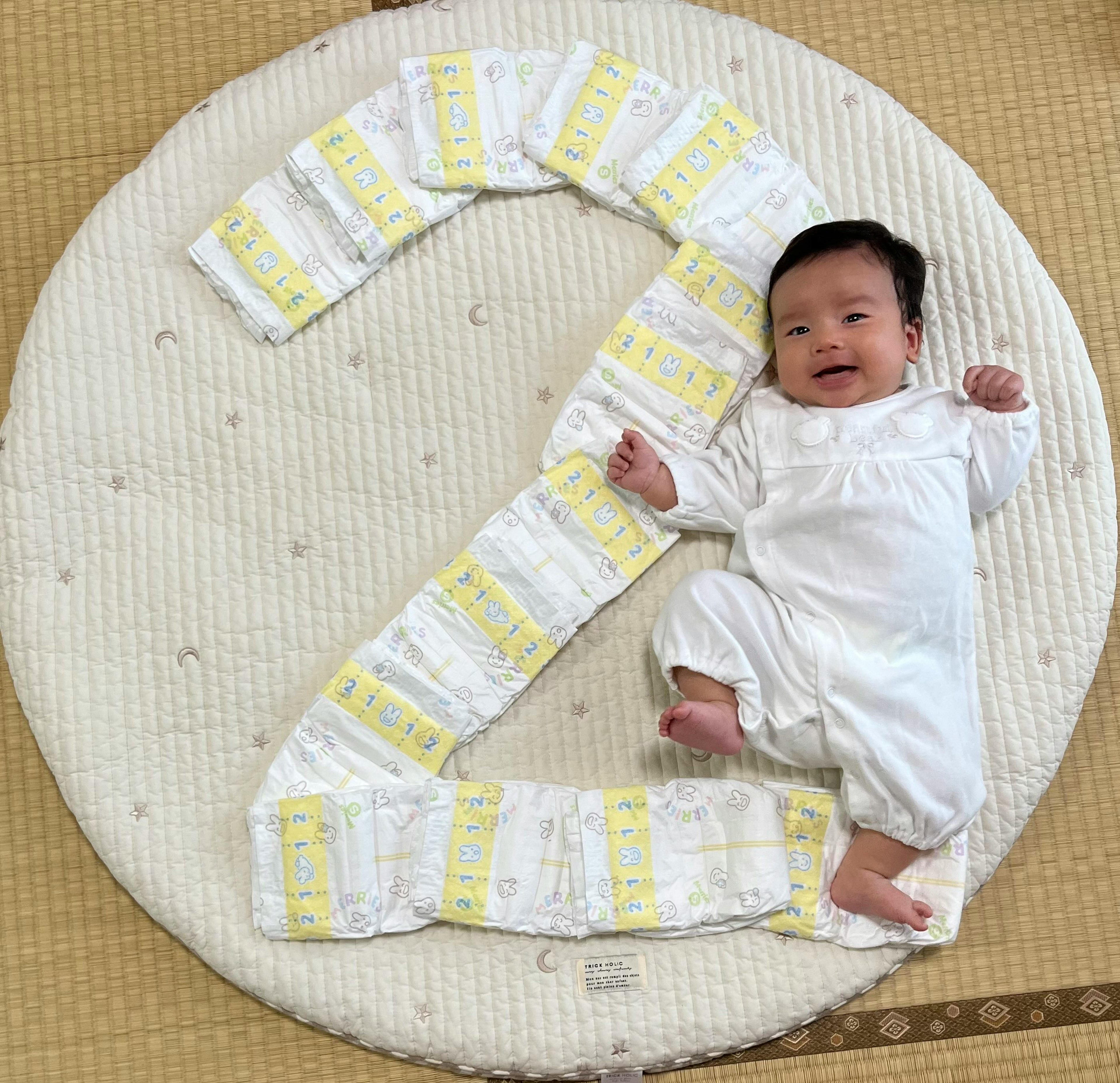 A baby in white clothing lying on a round mat surrounded by diapers arranged in the shape of the number 2