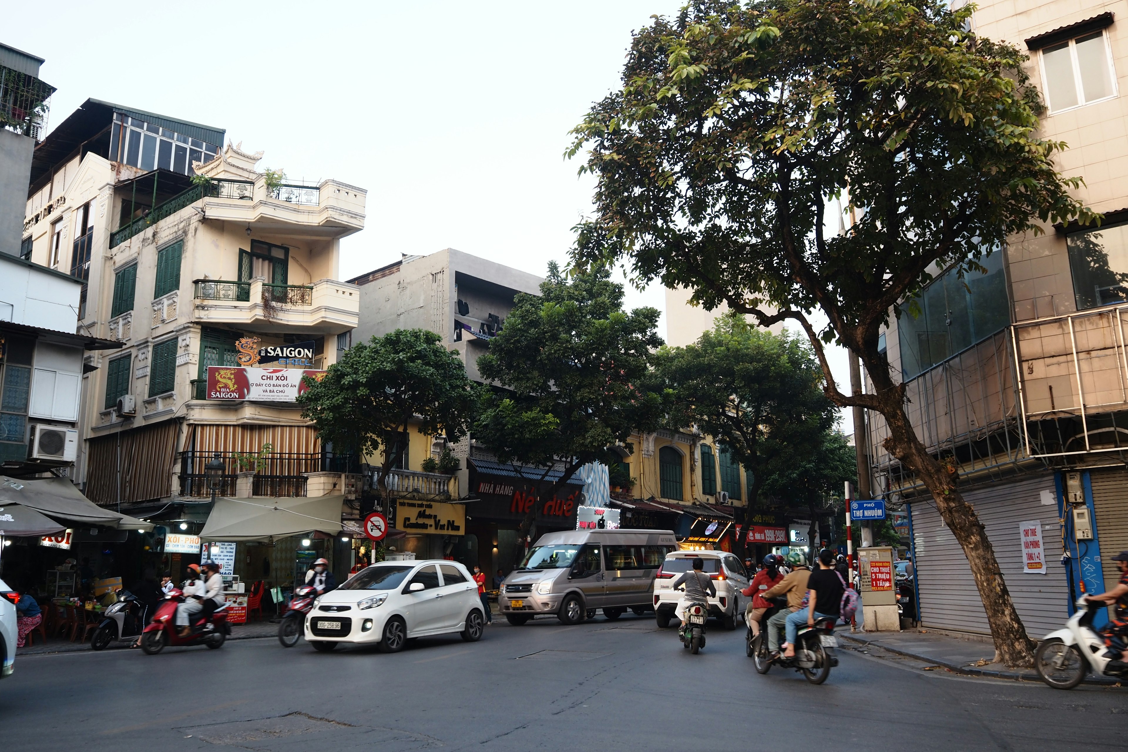 Scène de rue à Hanoi avec des voitures et des motos passant sous des arbres luxuriants