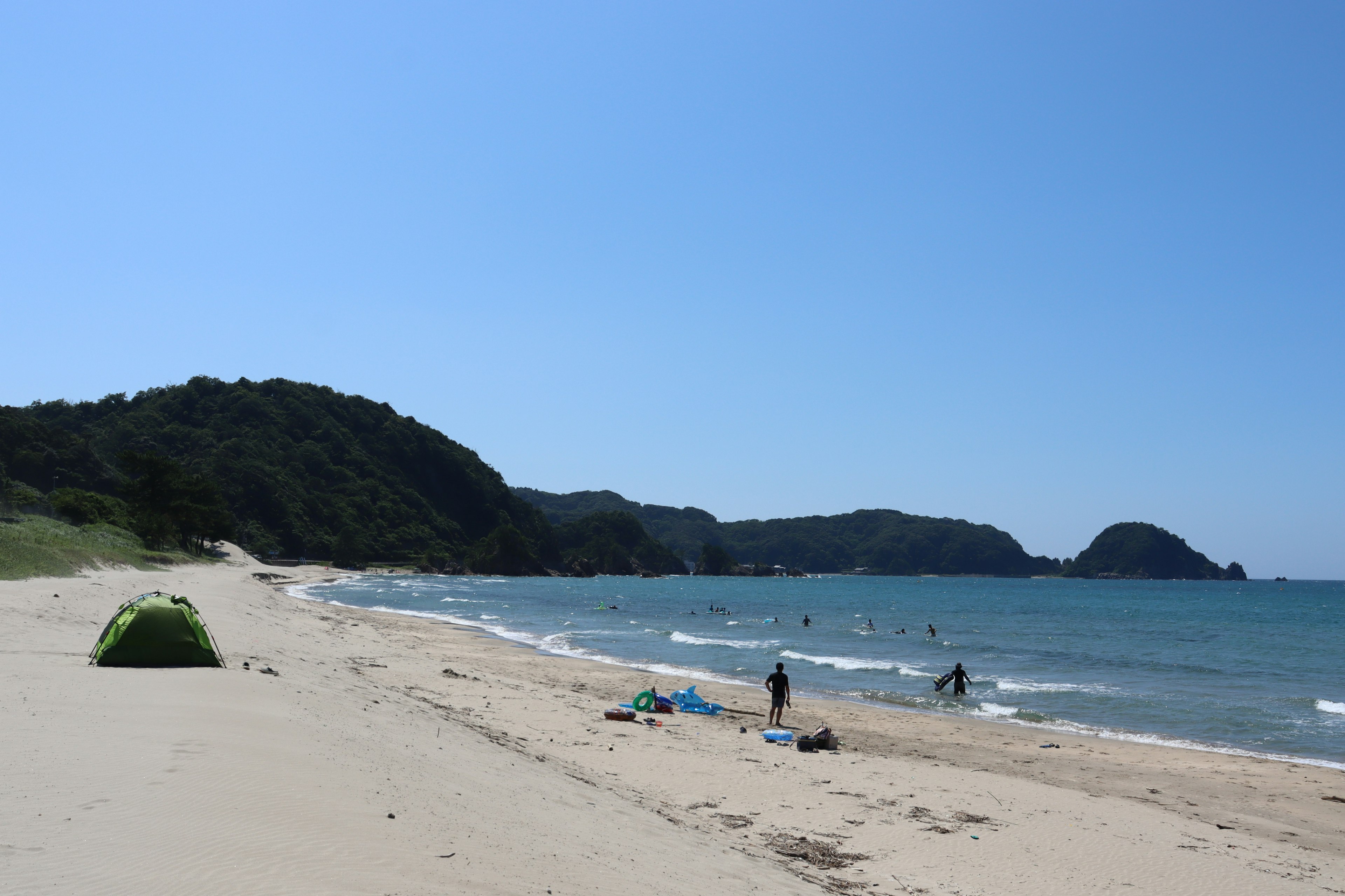 ทิวทัศน์ชายหาดที่สวยงามพร้อมมหาสมุทรสีน้ำเงินและชายฝั่งทรายที่มีผู้คนเพลิดเพลินกับน้ำและเต็นท์สีเขียว