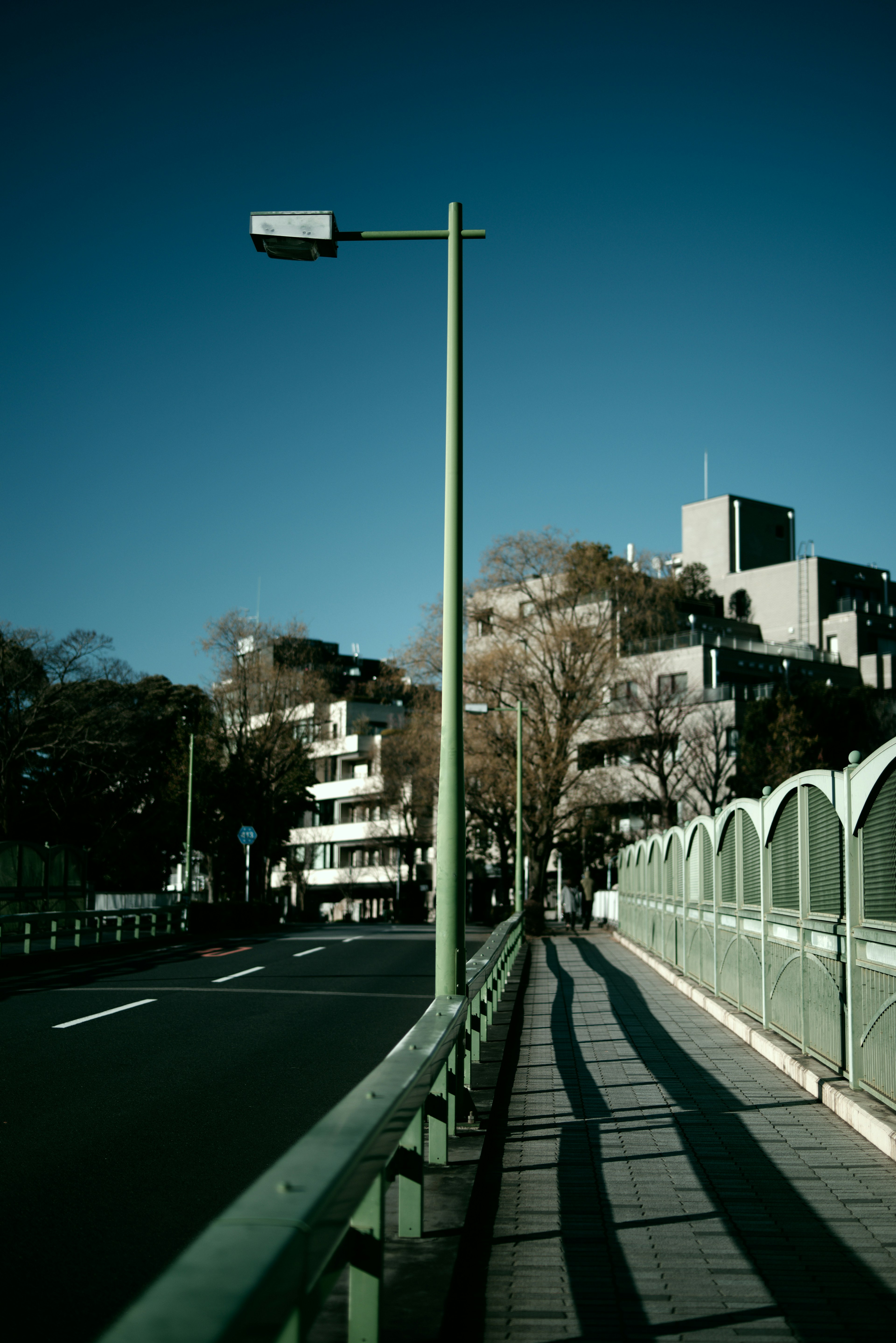 Lampione verde sotto un cielo blu con vista su un marciapiede e parte di un ponte in un contesto urbano