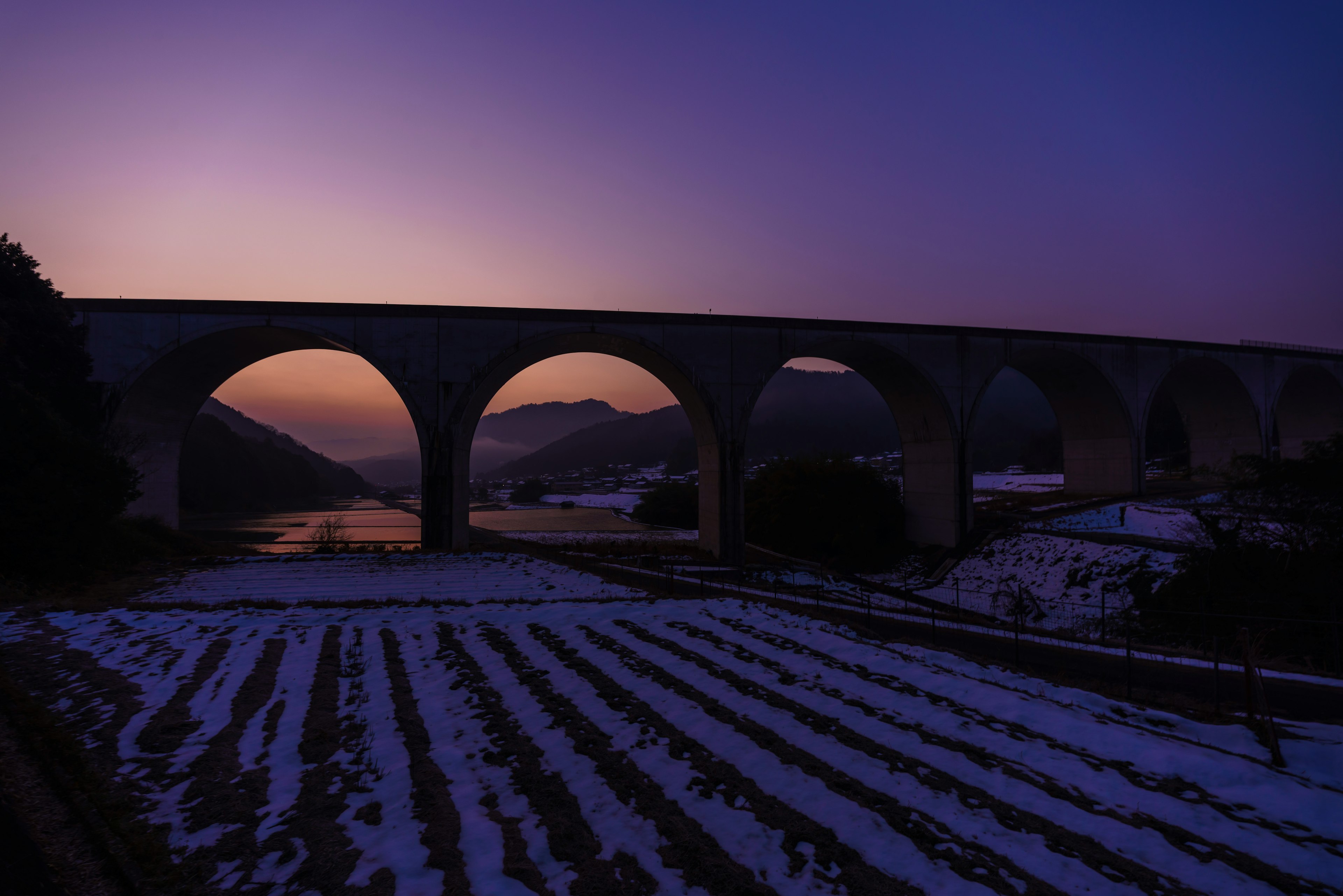 Large arch bridge under a purple sky with snow-covered farmland